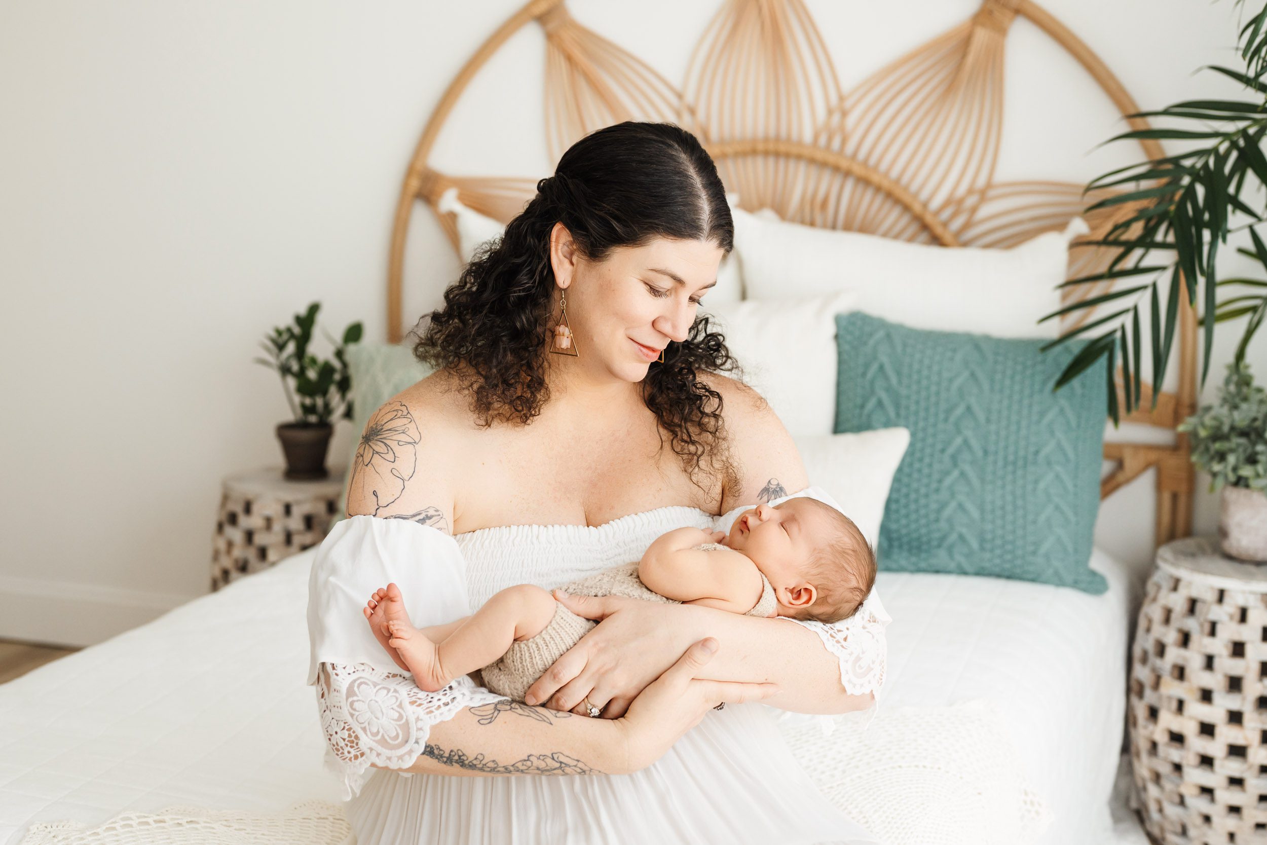 a new mom sitting on a bed cradling her baby boy in her arms and smiling down at him during a newborn photoshoot