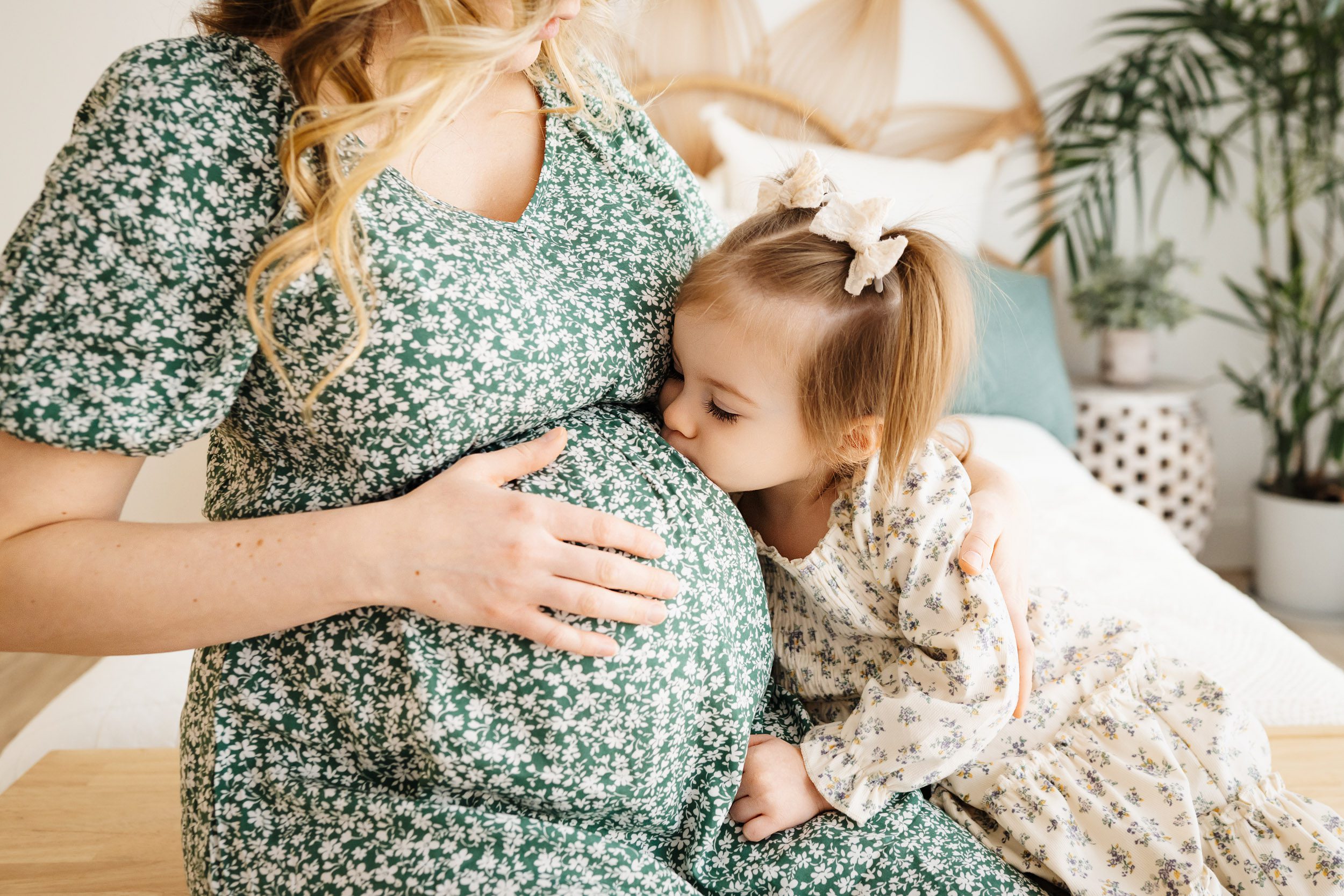 a close up picture of an expecting mom wearing a green and white floral dress touching her belly as her young daughter leans in and gives her belly a kiss during a warm & cozy maternity photoshoot