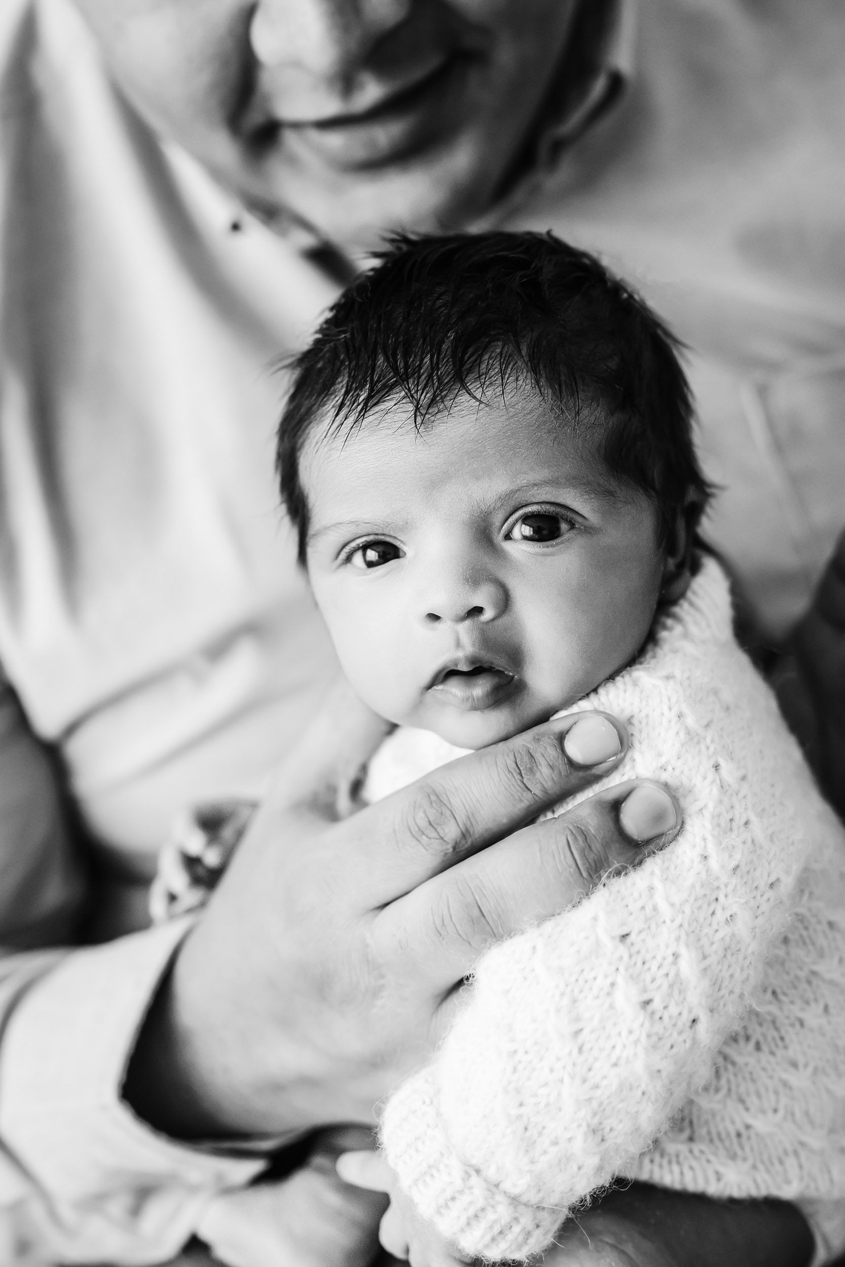 a black and white photo of a dad holding his baby girl in her arms and burping her as the baby gazes intently into the camera during a family newborn photoshoot