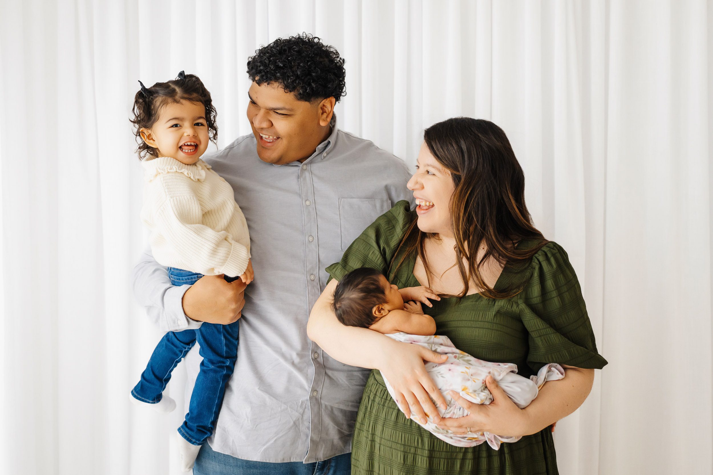 a family of four standing in front of a wall of white curtains as dad holds his older daughter in his arms and mom cradles her newborn baby in her arms during a family newborn photoshoot