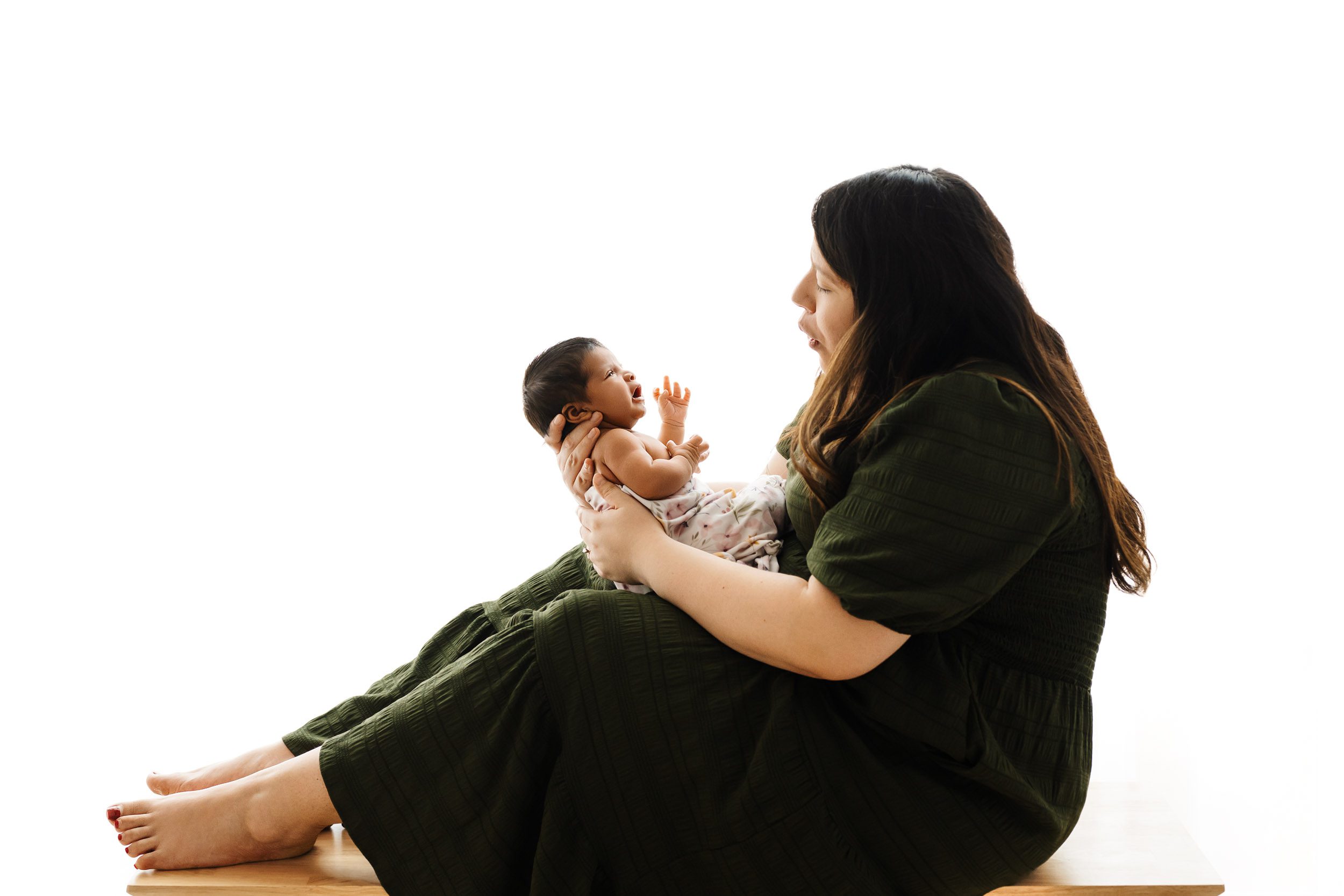 a backlit photo of a new mom sitting on a bench holding her baby girl in her arms and smiling down at her during a family newborn photoshoot