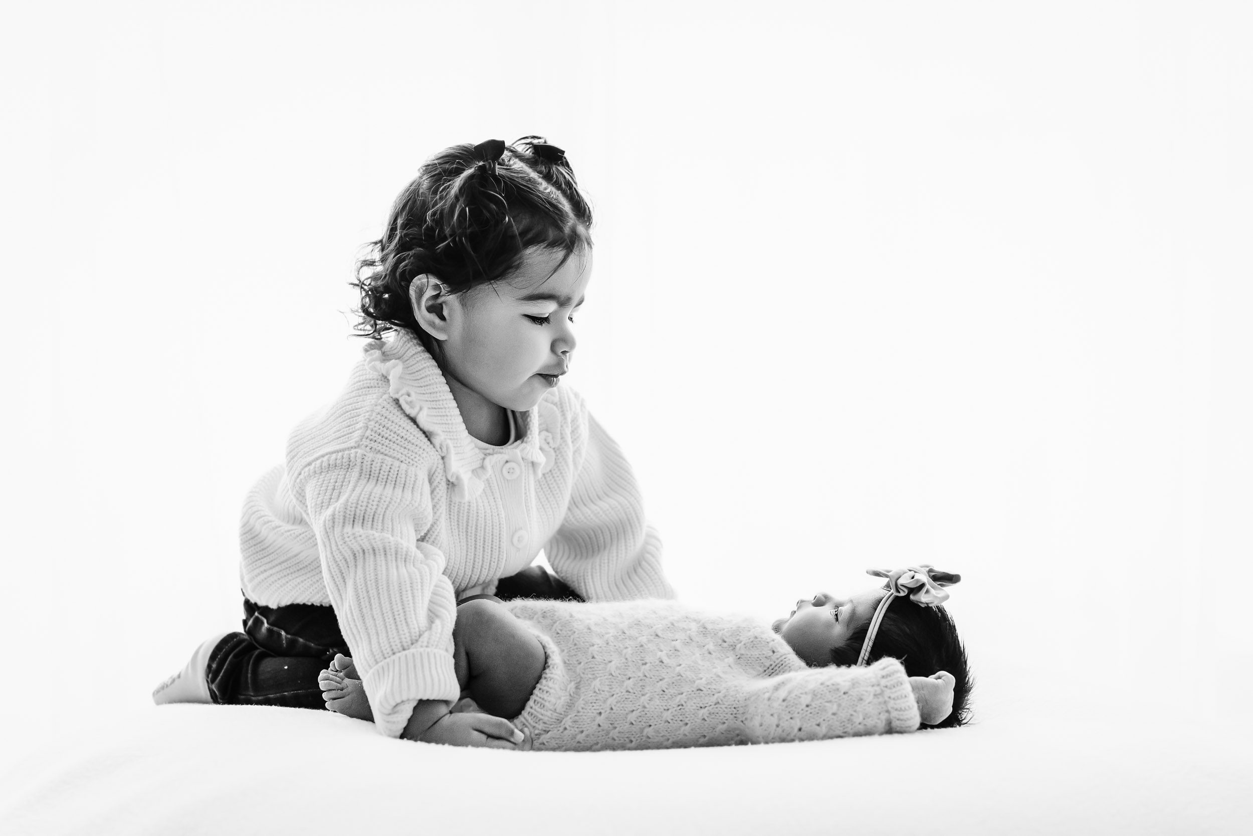 a black and white backlit photo of a baby girl laying on a bean bag with her sister kneeling next to her and smiling down at her during a family newborn photoshoot