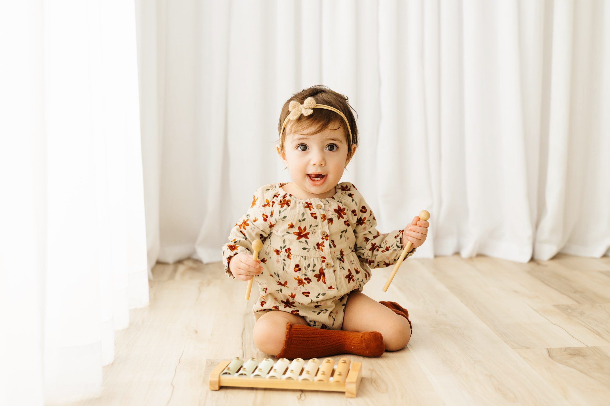 a young girl wearing a floral romper sitting on the floor and playing with a xylophone toy as she sticks her tongue out at the camera during a Pottstown 1st birthday photoshoot