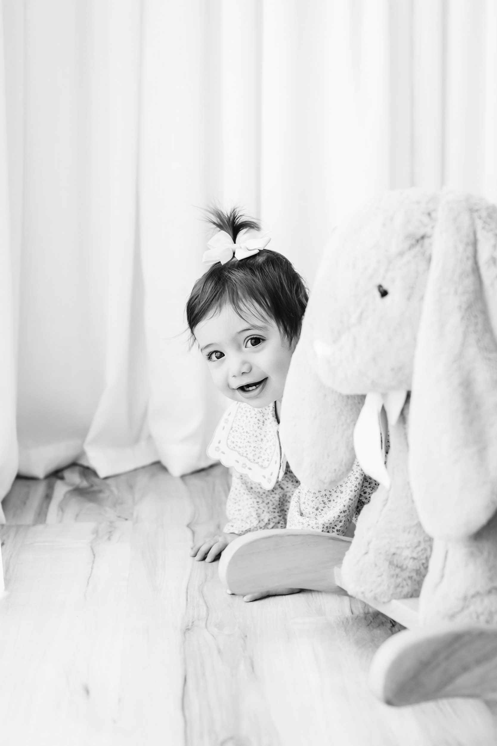 a black and white picture of a little girl peeking her head out from behind a bunny rocker toy and smiling at the camera during a Pottstown 1st birthday photoshoot