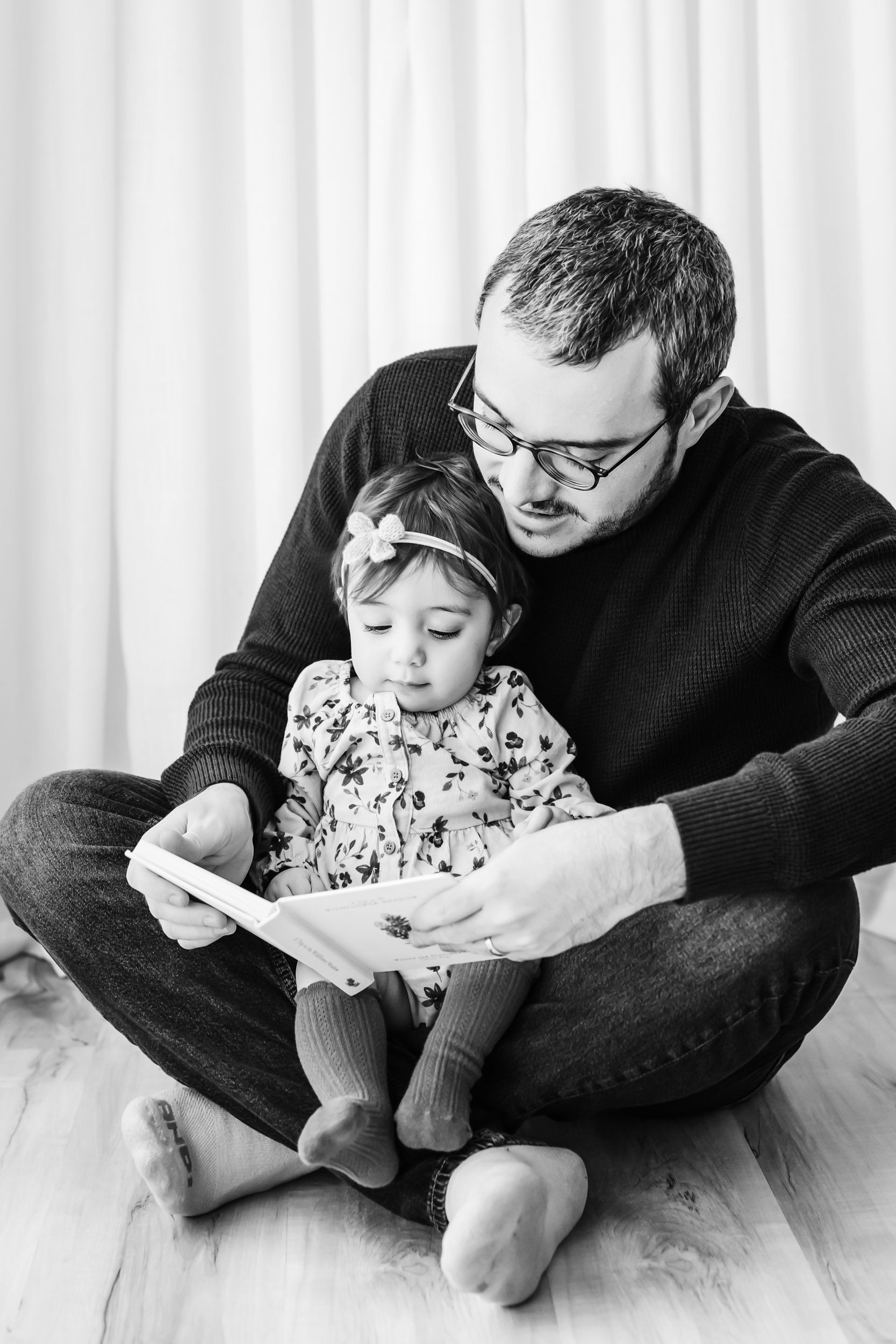 a black and white picture of a little girl sitting on her dad's lap as he reads her a story during a Pottstown 1st birthday photoshoot