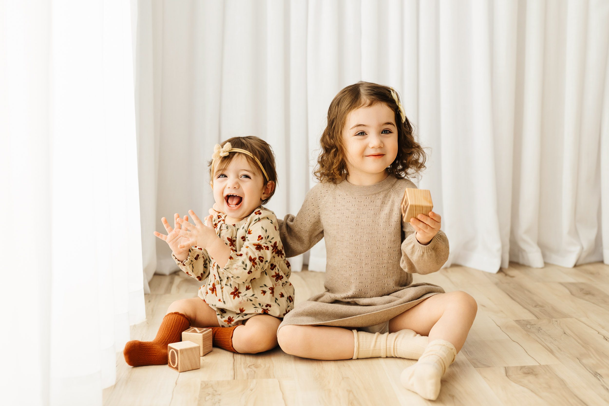 two young sisters sitting on the floor together playing with wooden blocks as they clap and smile at the camera during a Pottstown 1st birthday photoshoot