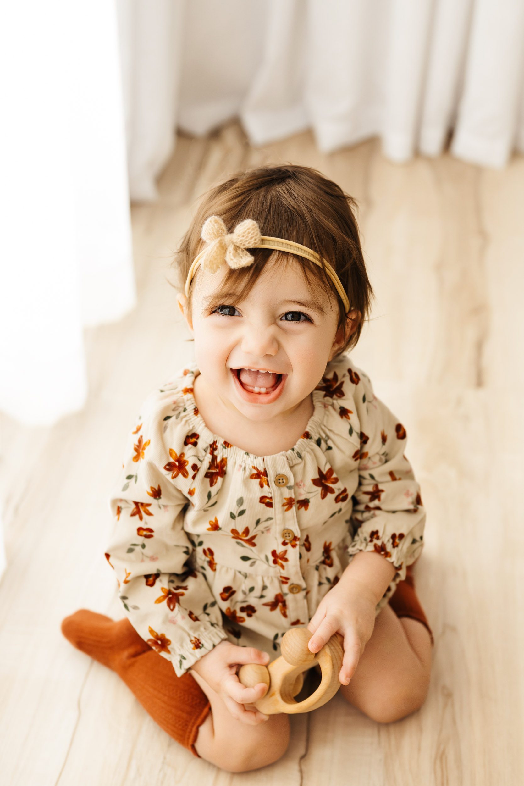 a picture taken from above of a little girl wearing a floral romper and holding a wooden toy car looking up with a huge smile on her face during a Pottstown 1st birthday photoshoot
