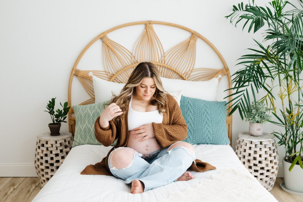 a pregnant mom wearing a chunky knit cardigan over a white bra and jeans sitting on a bed and gently touching her belly during a bare belly maternity photoshoot