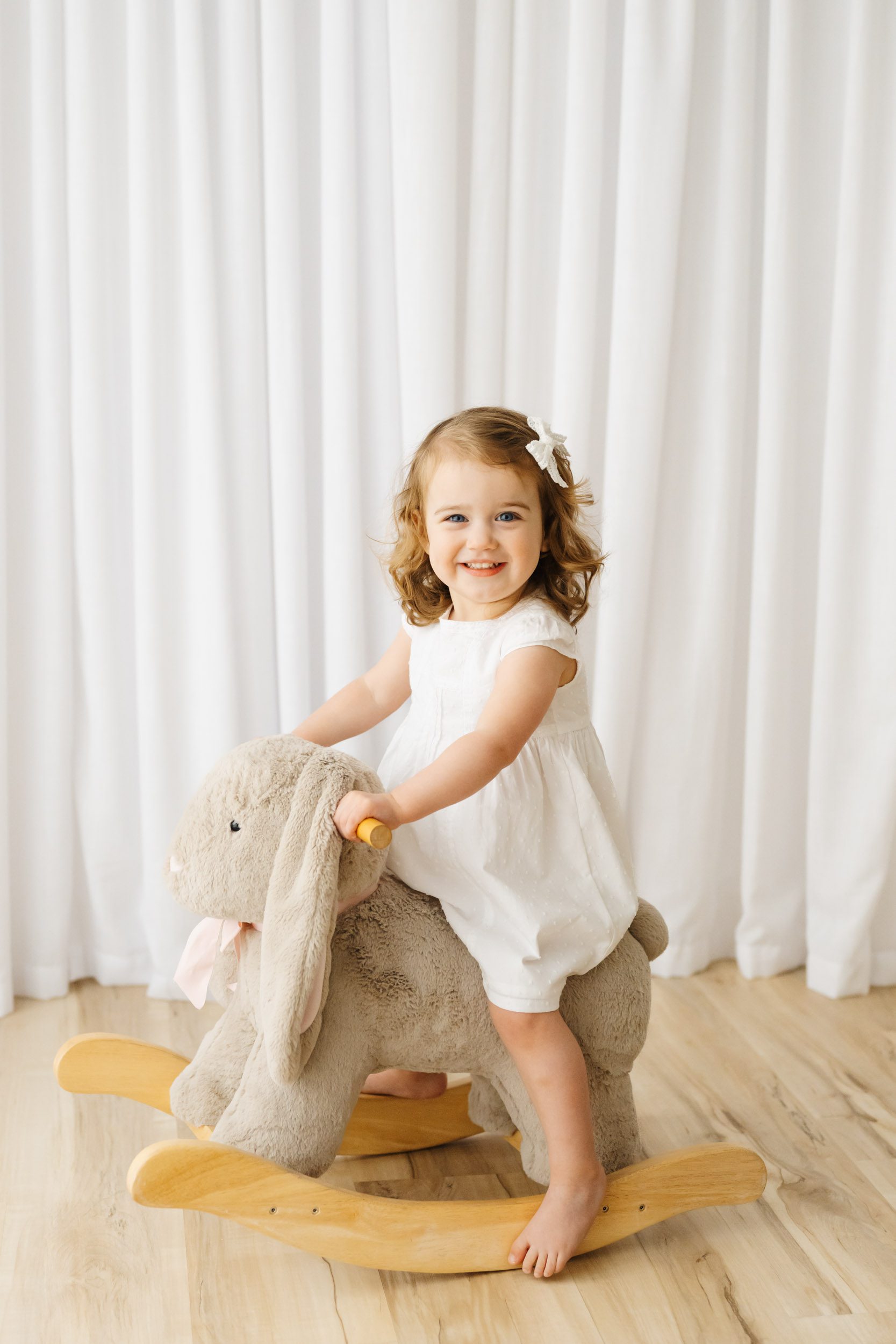 a young girl riding on a bunny rocker toy and smiling at the camera during a 2nd birthday photoshoot