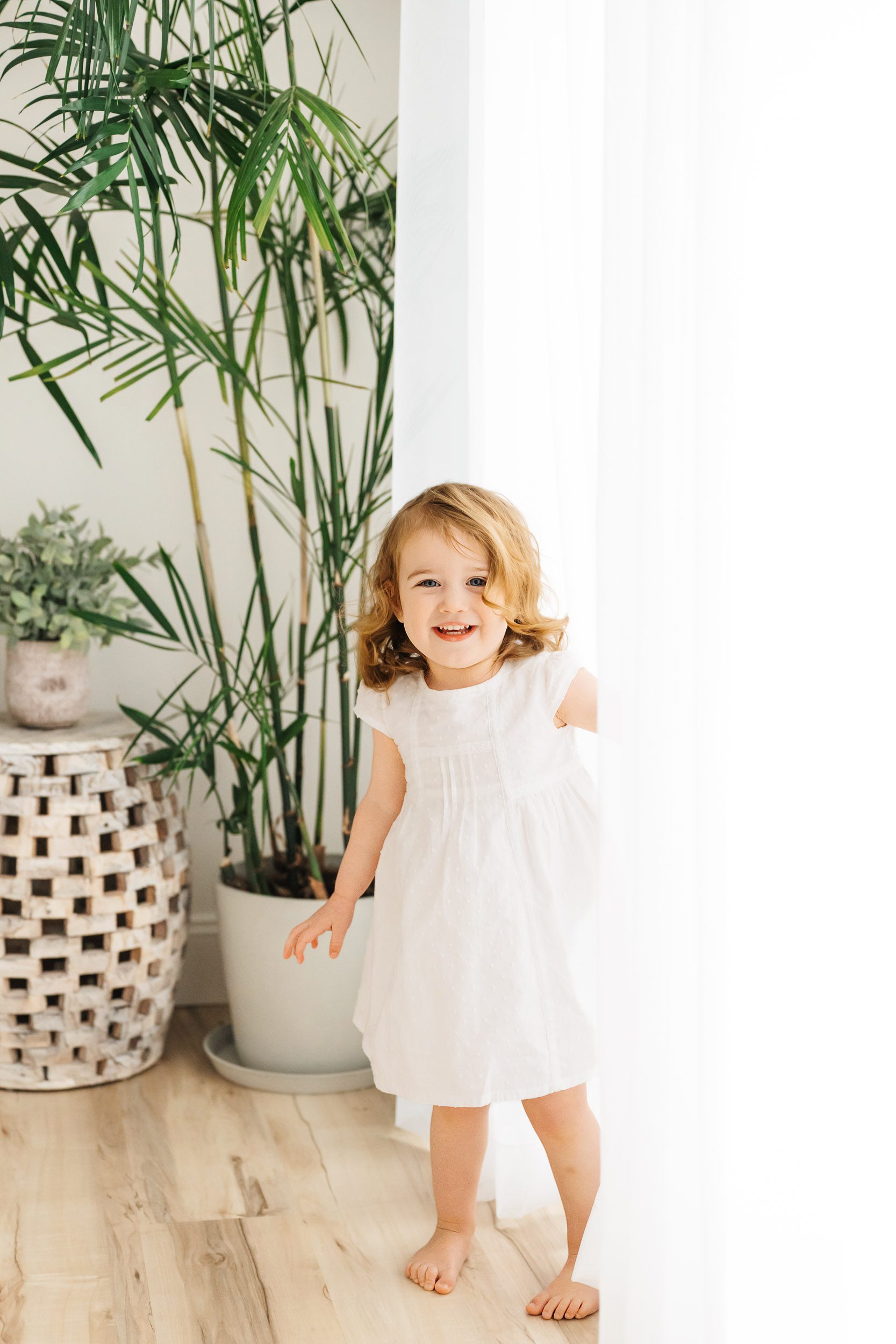 a young girl standing next to a wall of white curtains and smiling at the camera during a 2nd birthday photoshoot