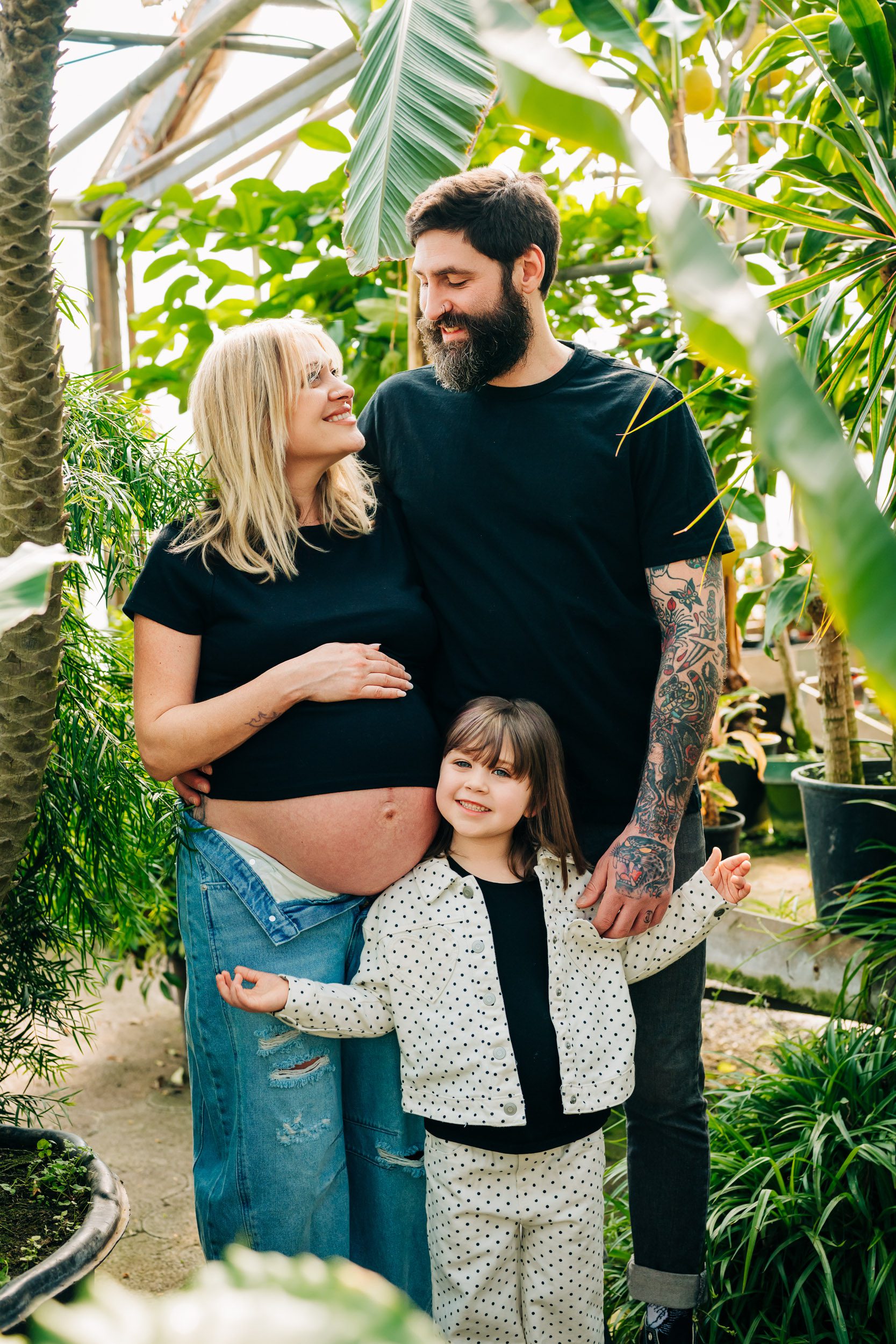 a family of three standing in a greenhouse surrounded by foliage as mom rests her hand on her belly and smiles at dad while their young daughter stands in front of them smiling directly at the camera during a greenhouse maternity photoshoot