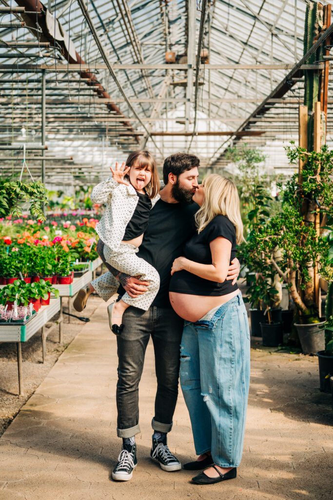 an expecting mom wearing jeans and a fitted black crop top kissing dad as he holds their young daughter on his hip and she sticks her tongue out at the camera during a greenhouse maternity photoshoot