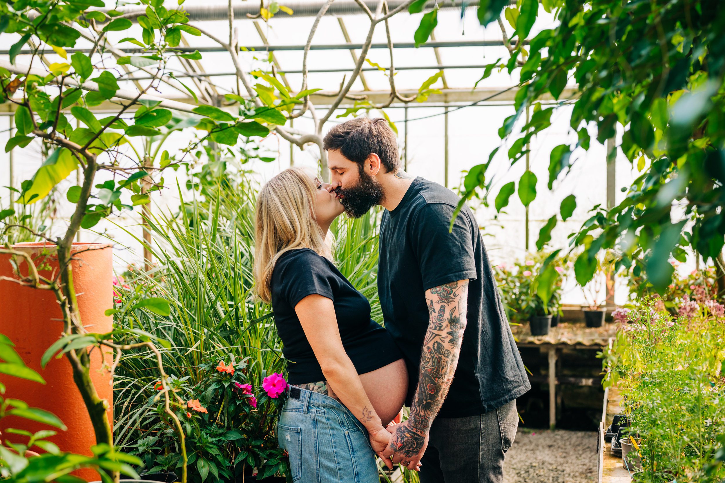 an expecting mom wearing jeans and a black crop top standing in the middle of greenery and foliage as she kisses her husband during a greenhouse maternity photoshoot