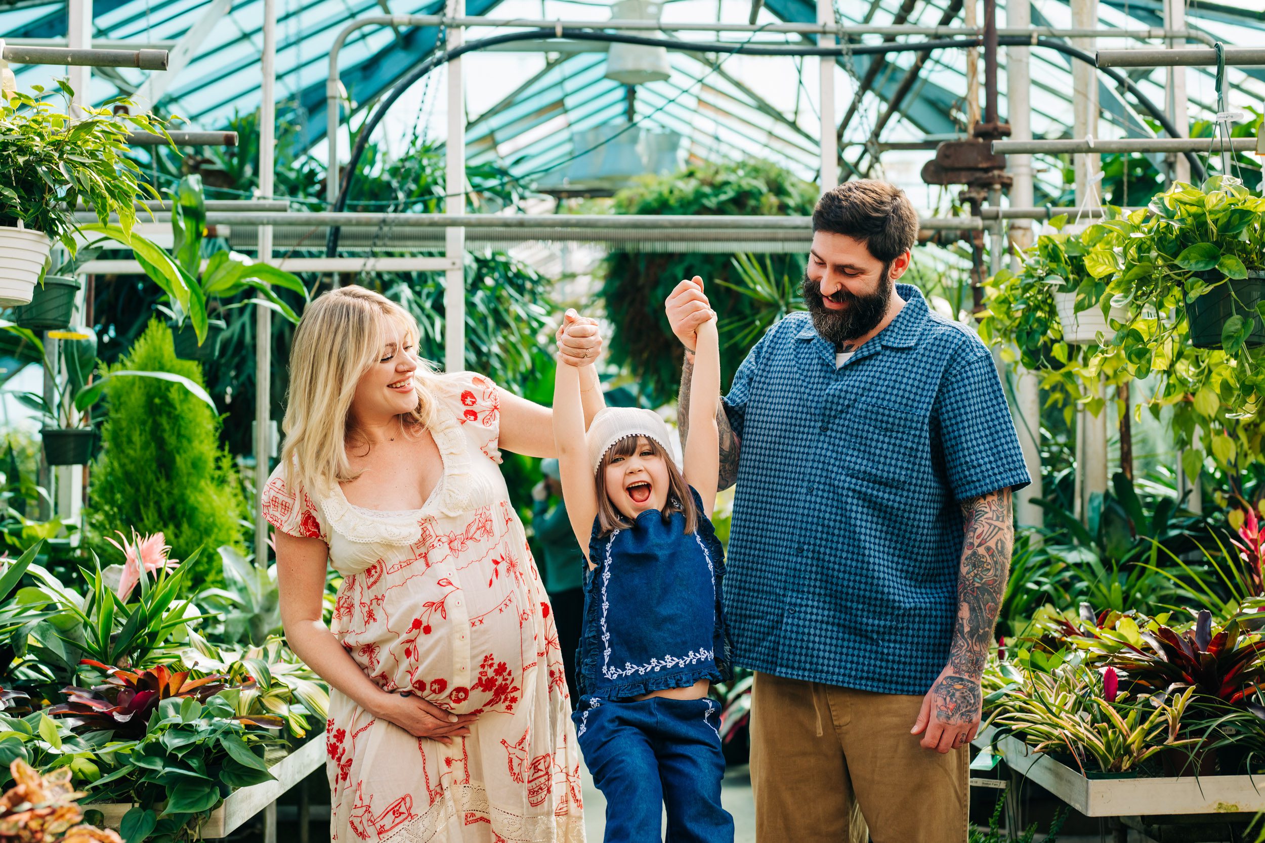 a mom & dad holding their young daughter's hands and lifting her up in the air as they all laugh during a greenhouse maternity photoshoot