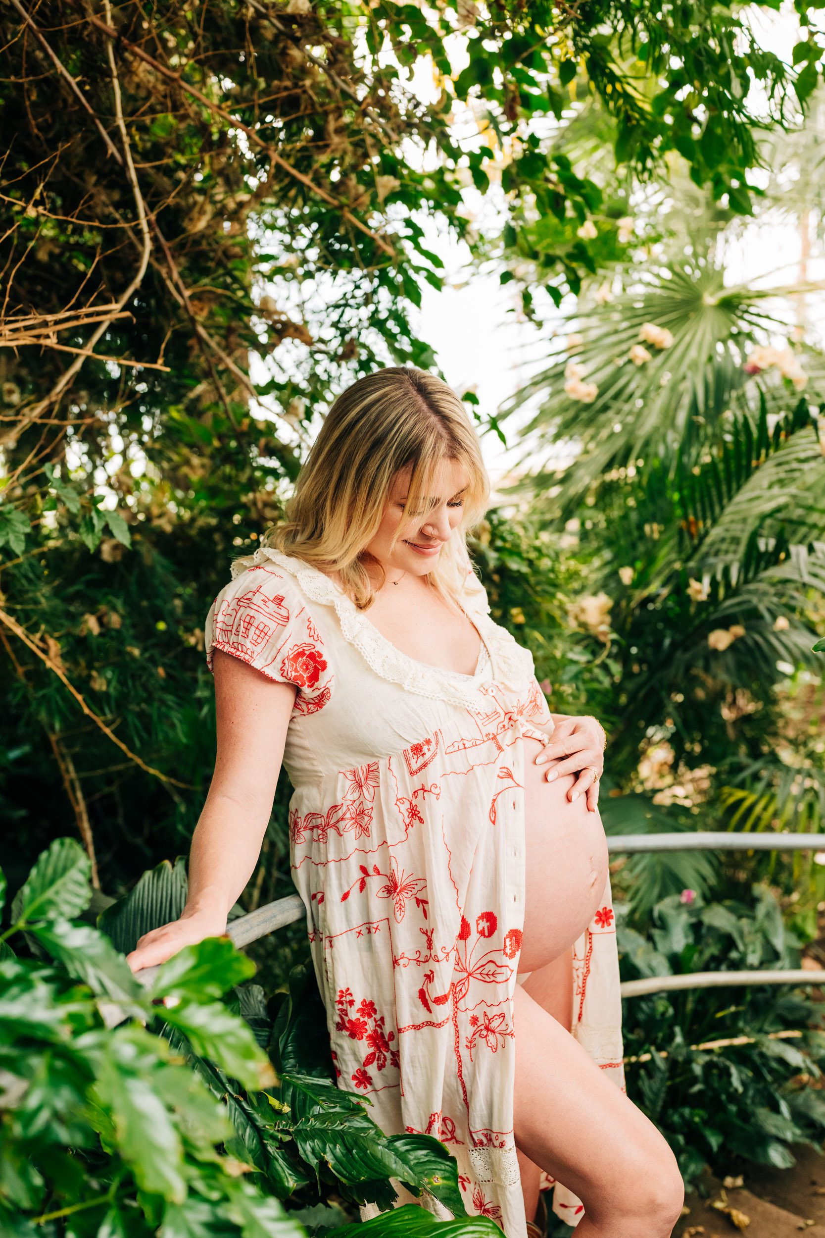 an expecting mom wearing a white dress with red flowers on it standing on a curved staircase surrounded by lush greenery all around her as she smiles down at her belly during a greenhouse maternity photoshoot