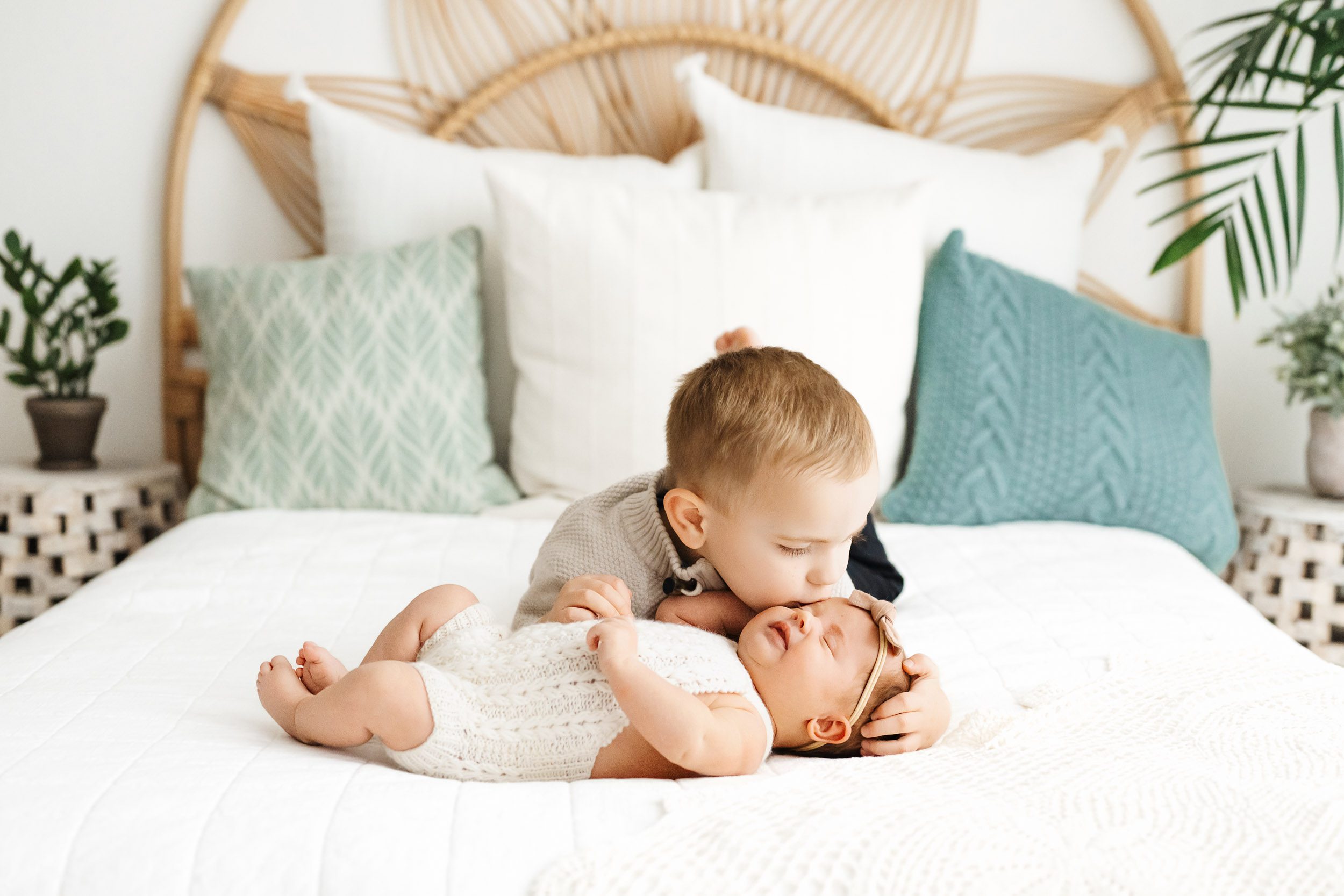a big brother laying on a bed with his baby sister and gently giving her a kiss on the head during a newborn photoshoot with siblings