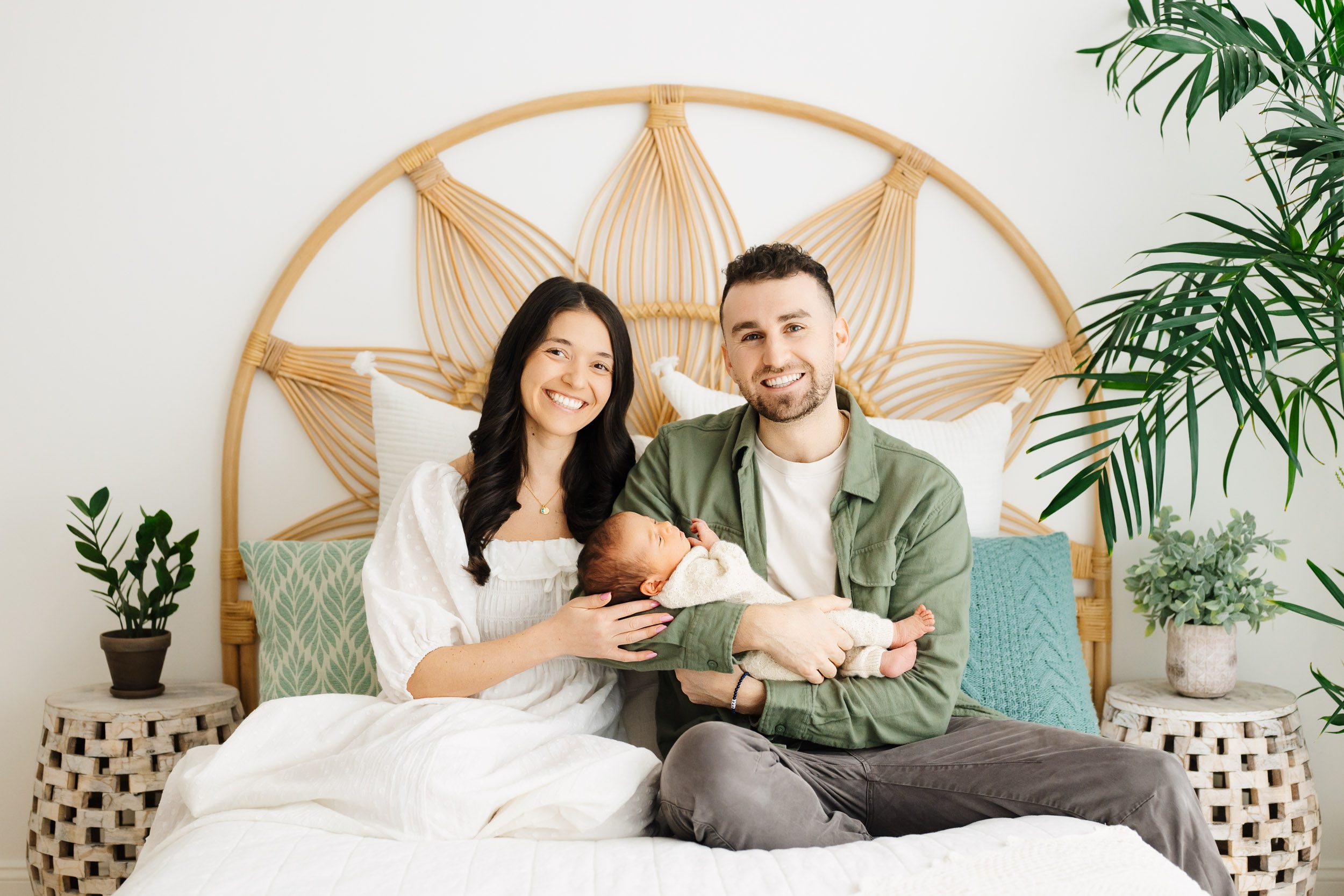 new parents sitting on a bed with their baby boy snuggled in their arms and smiling at the camera during an organic newborn photography session