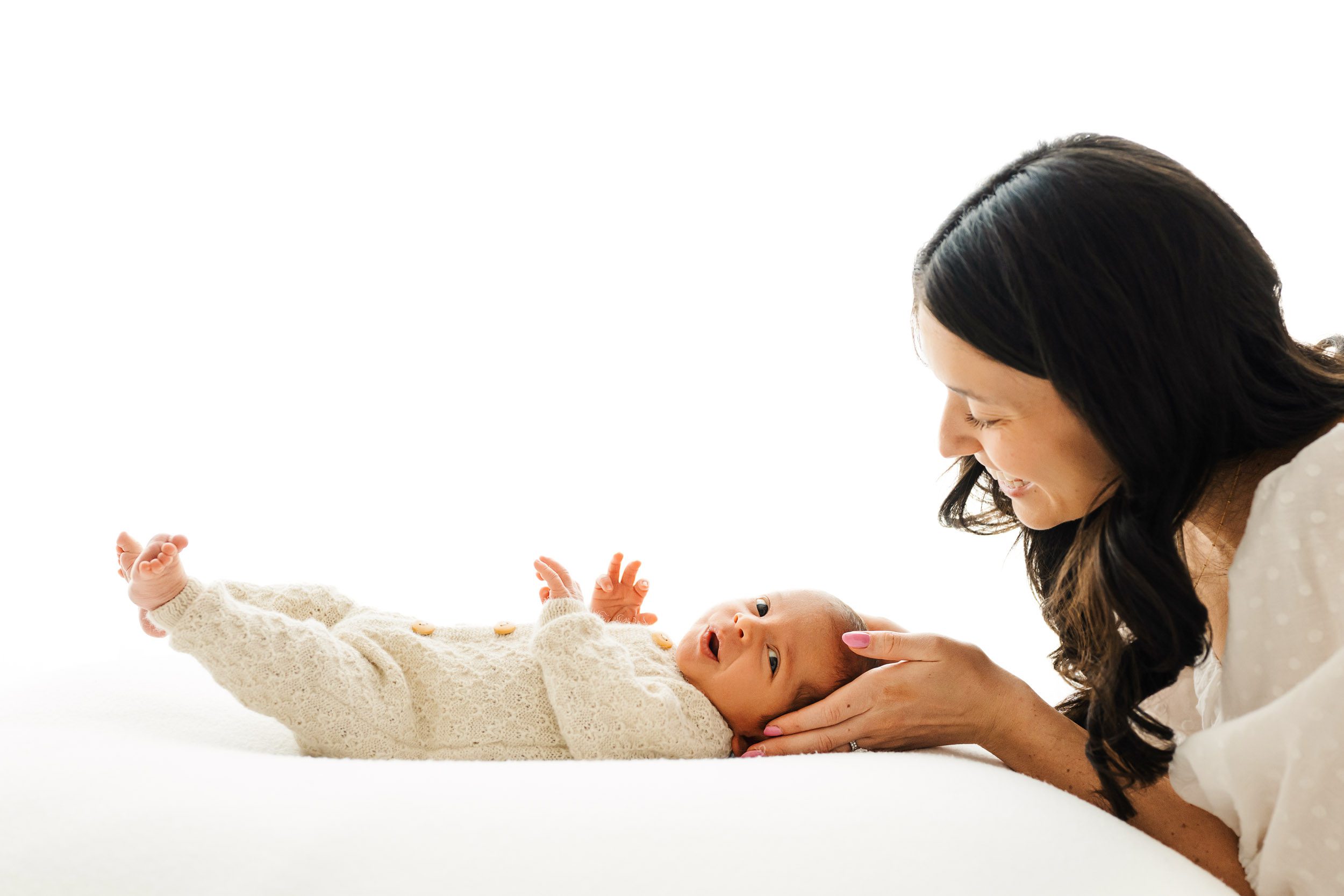 a backlit photo of a baby boy laying on a bean bag and looking to the side with a smirk on his face as his mom holds his head in her hands and smiles down at him during an organic newborn photography session