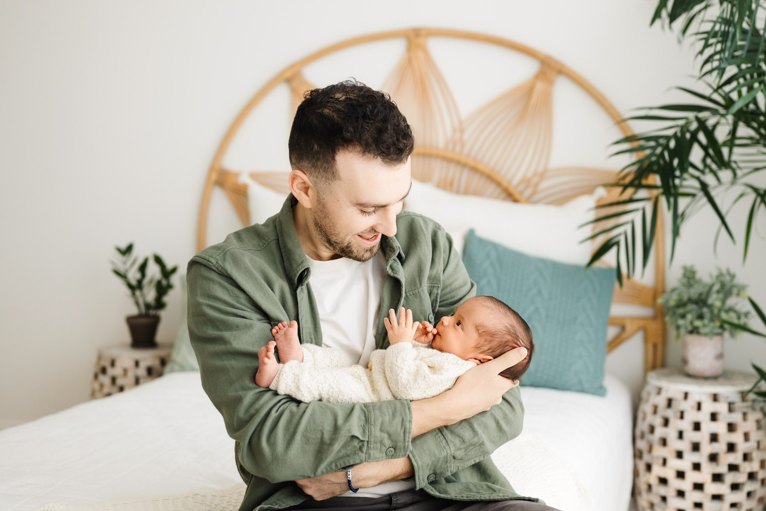 a new dad sitting on a bed and cradling his baby boy in his arms as he smiles down at him during an organic newborn photography session