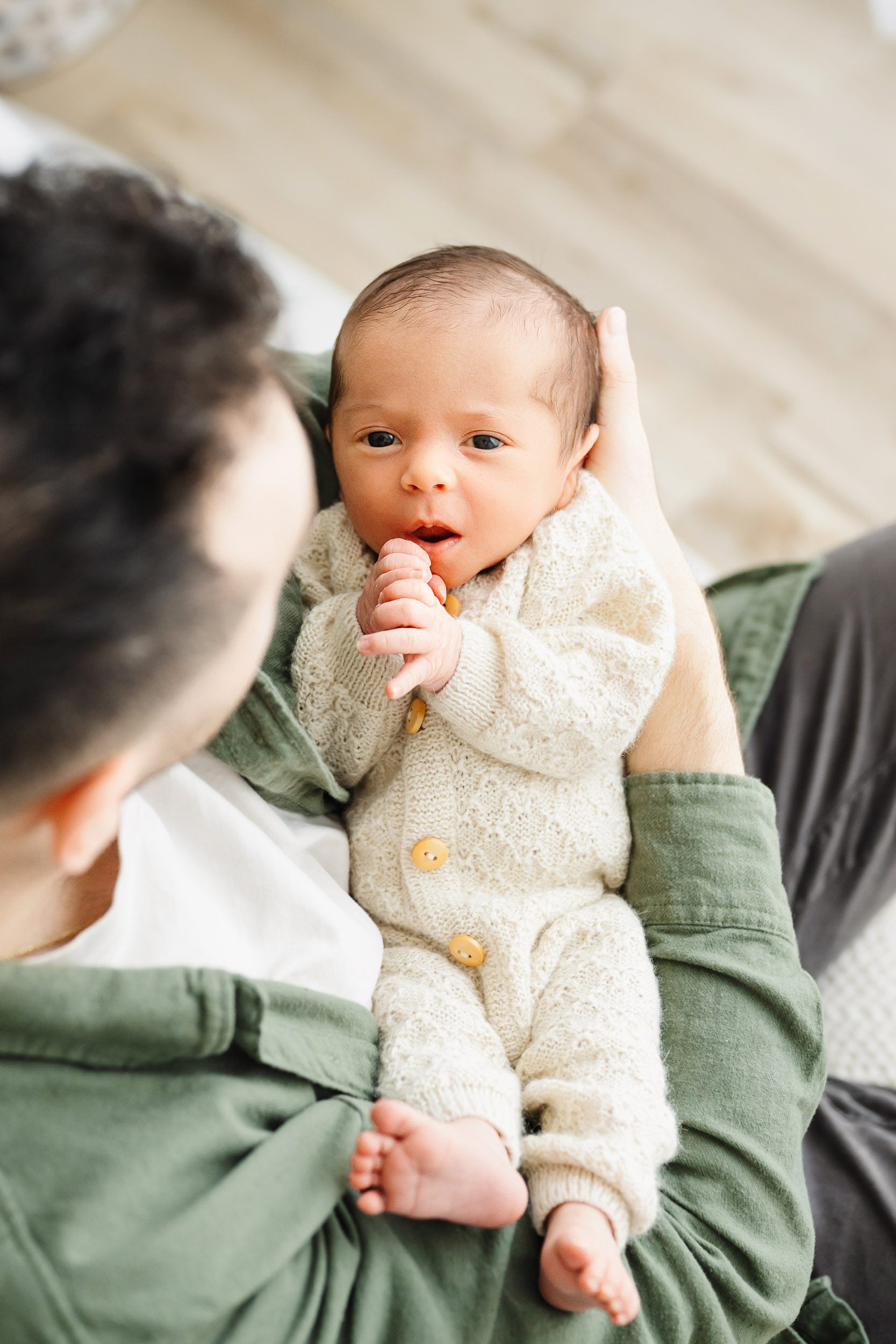 a photo taken from above of a baby boy cradled in his dad's arms and touching his hands to his mouth as he gazes up at the camera during an organic newborn photography session