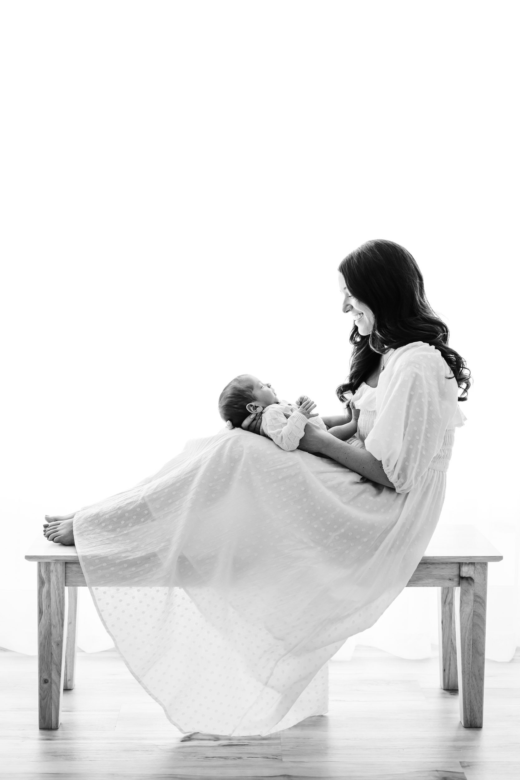 a black and white backlit photo of a new mom sitting on a bench with her baby boy resting on her legs as she smiles down at him during an organic newborn photography session