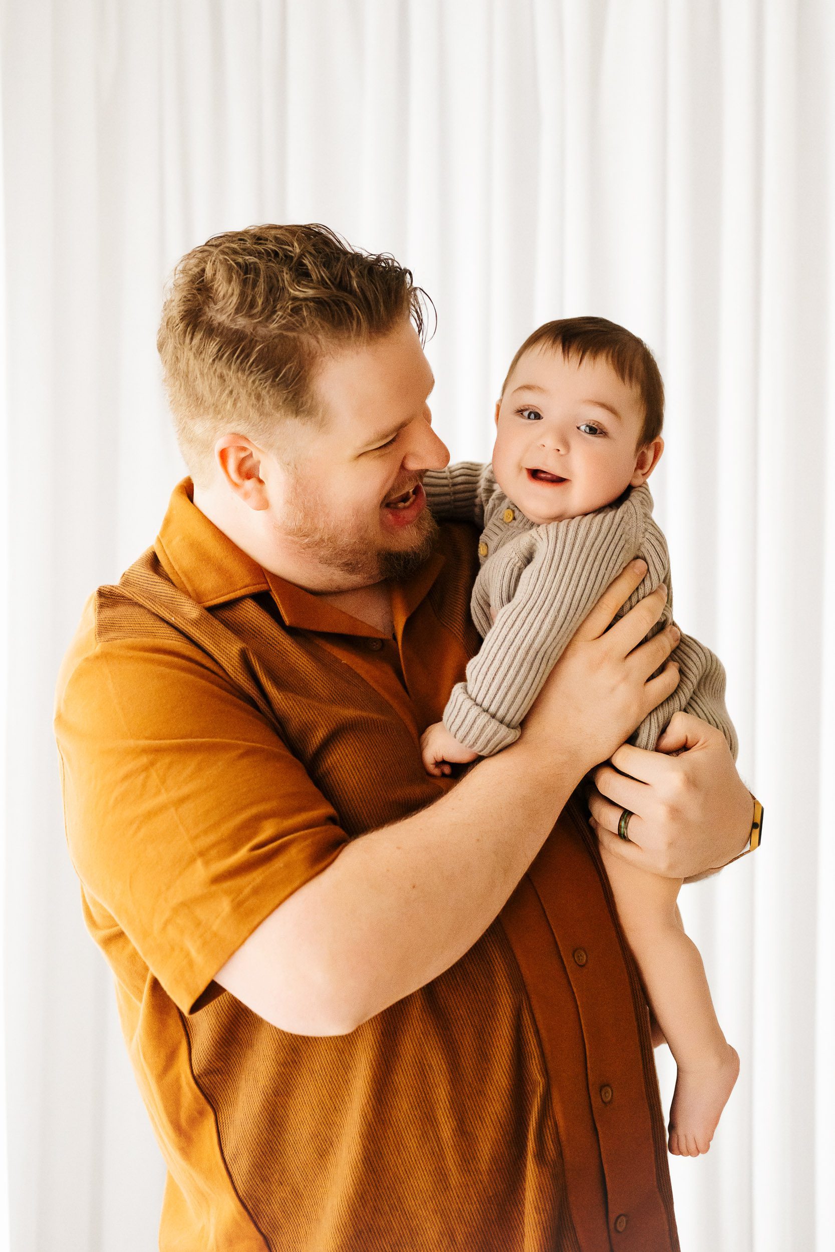 a dad holding his young son and smiling at him while the little boy smiles at the camera during a candid first birthday photoshoot