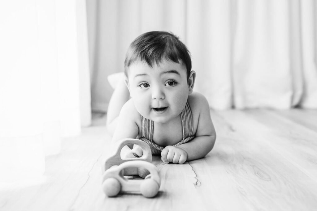 a black and white picture of a little boy laying on the floor and playing with two wooden toy cars during a candid first birthday photoshoot