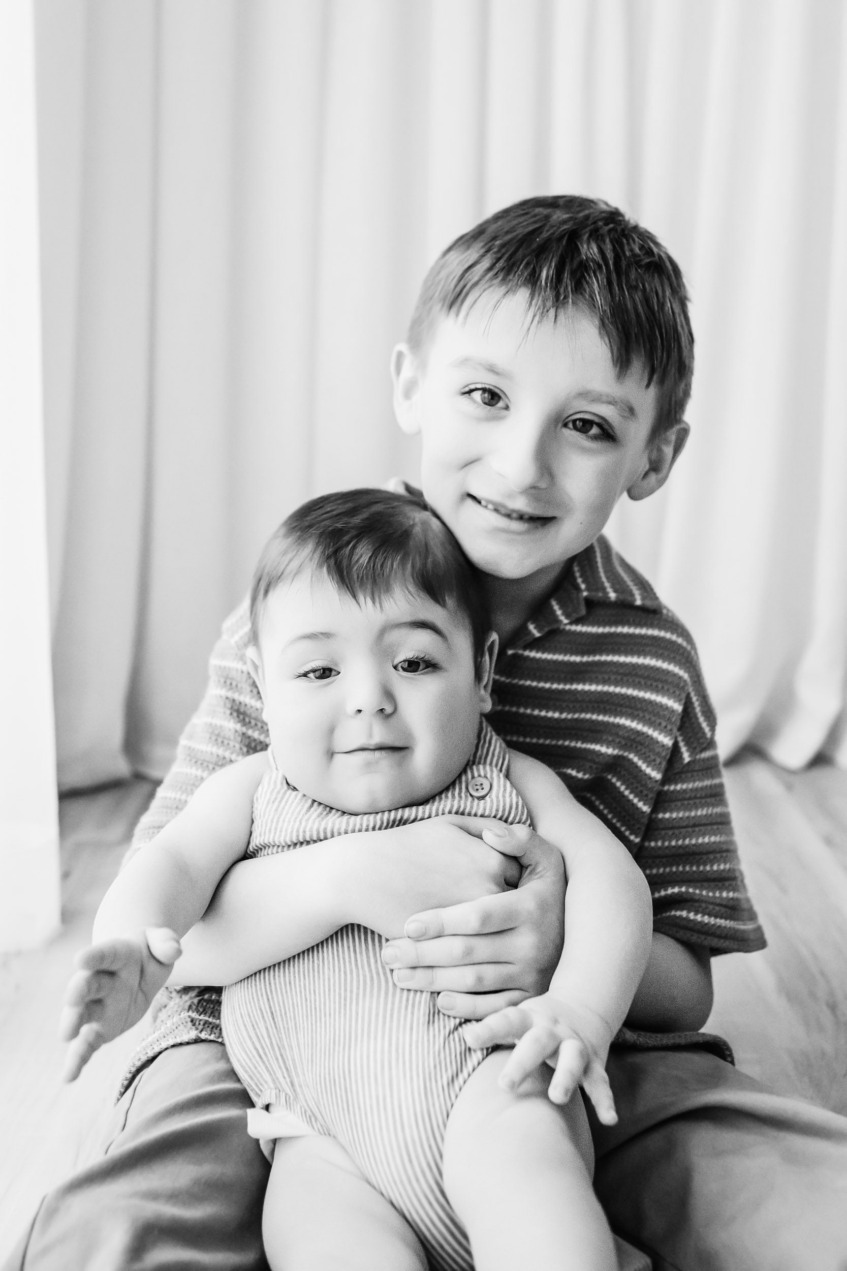 a black and white picture of an older brother sitting on the floor and hugging his younger brother as they both smile at the camera during a candid first birthday photoshoot