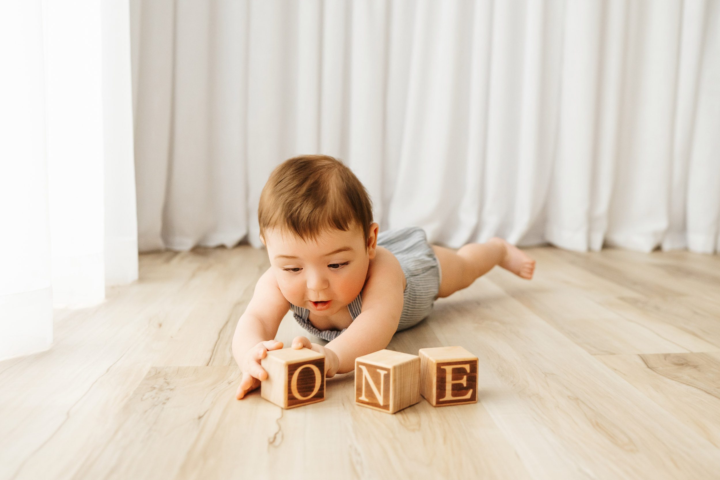a little boy laying on the floor with toy blocks that spell O N E laid out in front of him as he reaches out and touches the O block with his hands during a candid first birthday photoshoot