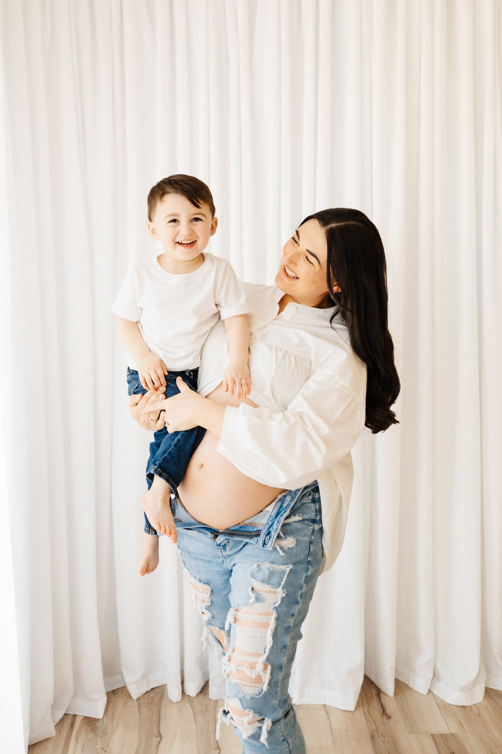 an expecting mom standing in front of a white curtain wall and holding her young son on her hip as she smiles at him while he smiles directly at the camera during a mommy & me maternity photoshoot