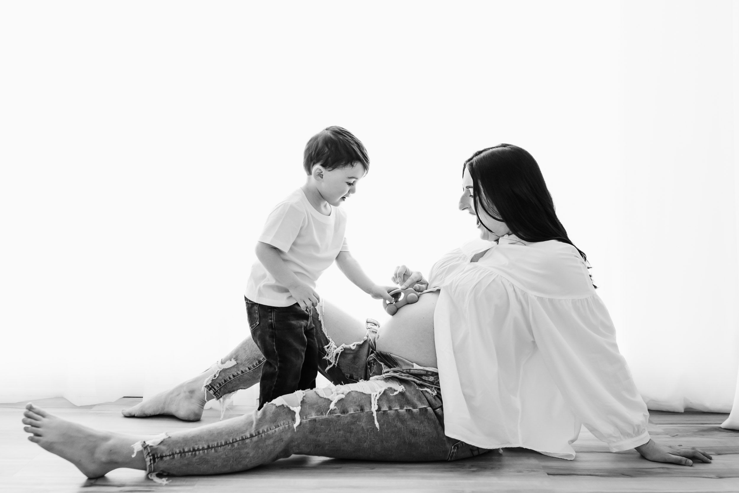 a black and white backlit photo of an expecting mom sitting on the floor wearing jeans and a white button up shirt with the buttons open to reveal her belly as her young son stands in front of her and drives his toy car up her belly during a mommy & me maternity photoshoot