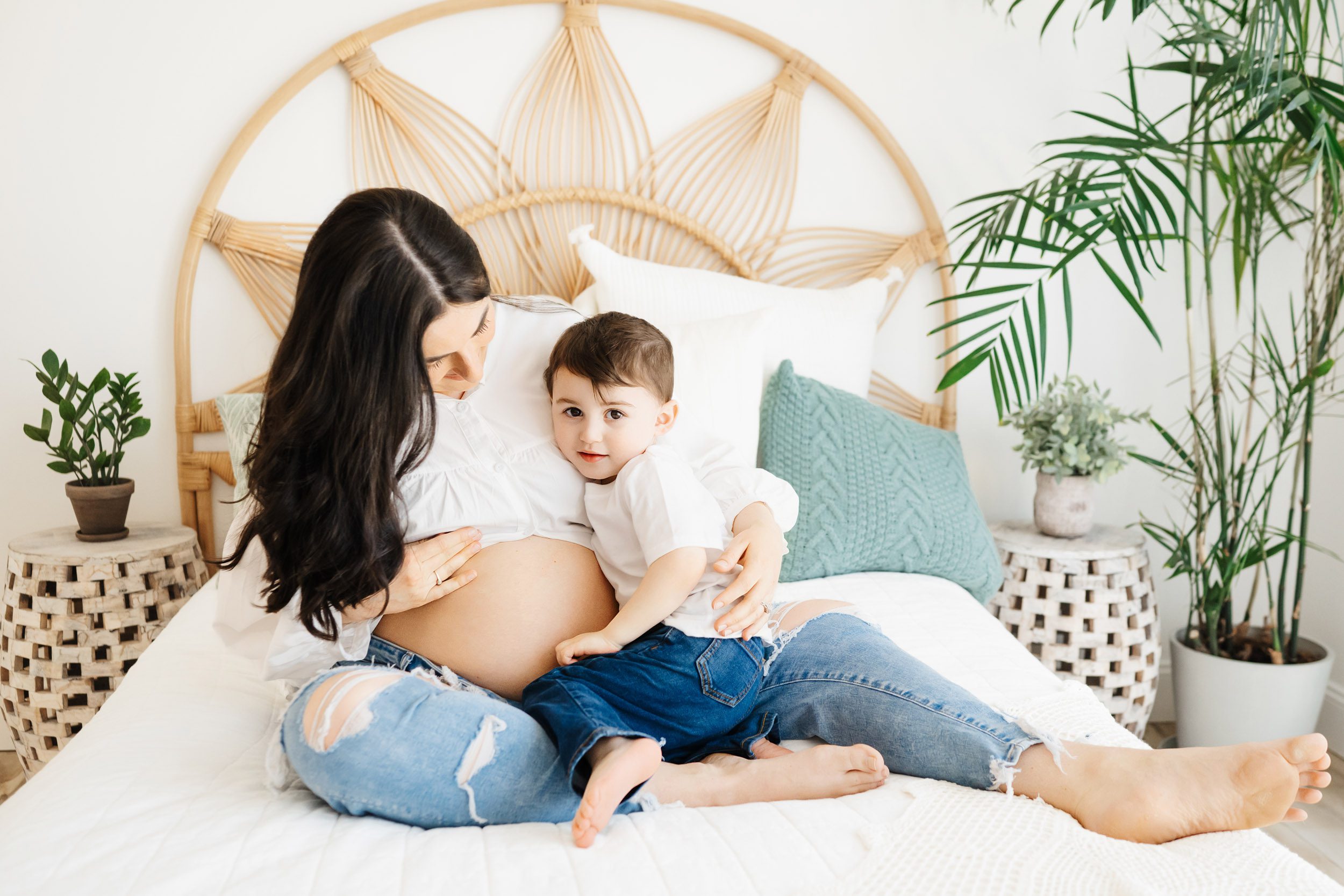 an expecting mother sitting on a bed with her young son snuggled on her lap hugging her as he gazes directly at the camera during a mommy & me maternity photoshoot