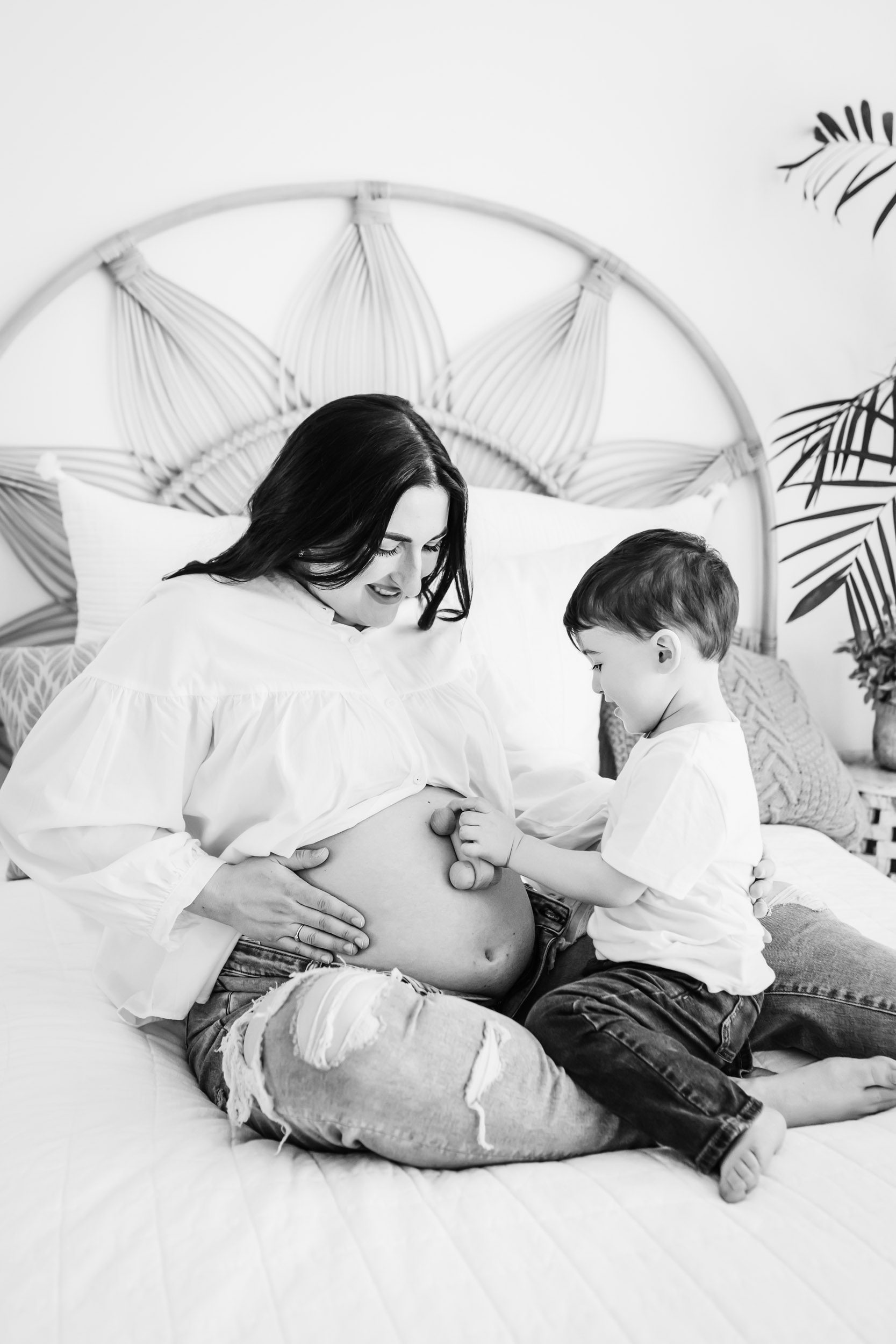 a black and white picture of a pregnant mom sitting on a bed with her young son as he drives a toy car up her belly during a mommy & me maternity photoshoot