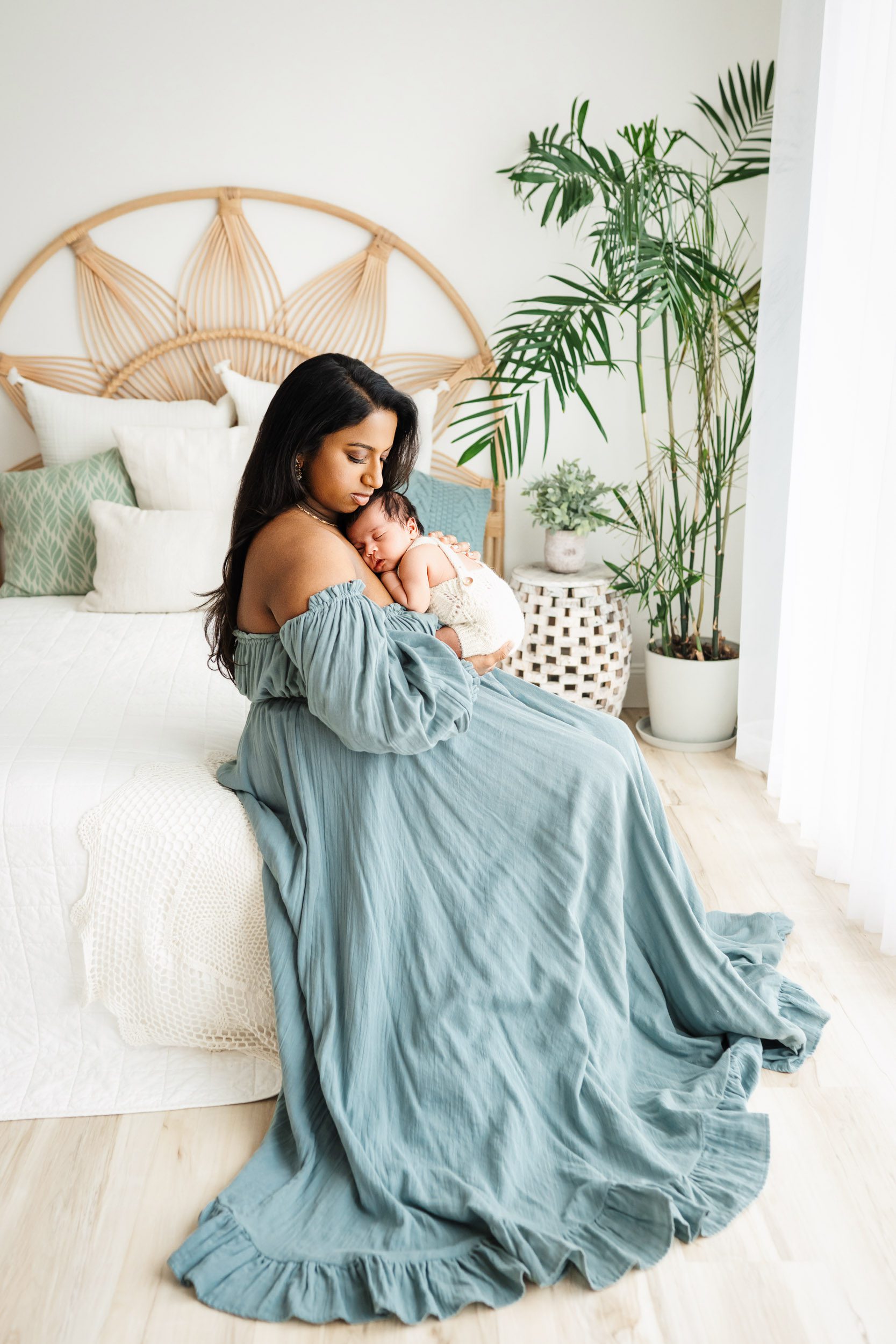 a new mom wearing a blue off the shoulder dress sitting on a bed with her baby boy snuggled up against her chest during a newborn photoshoot with mom