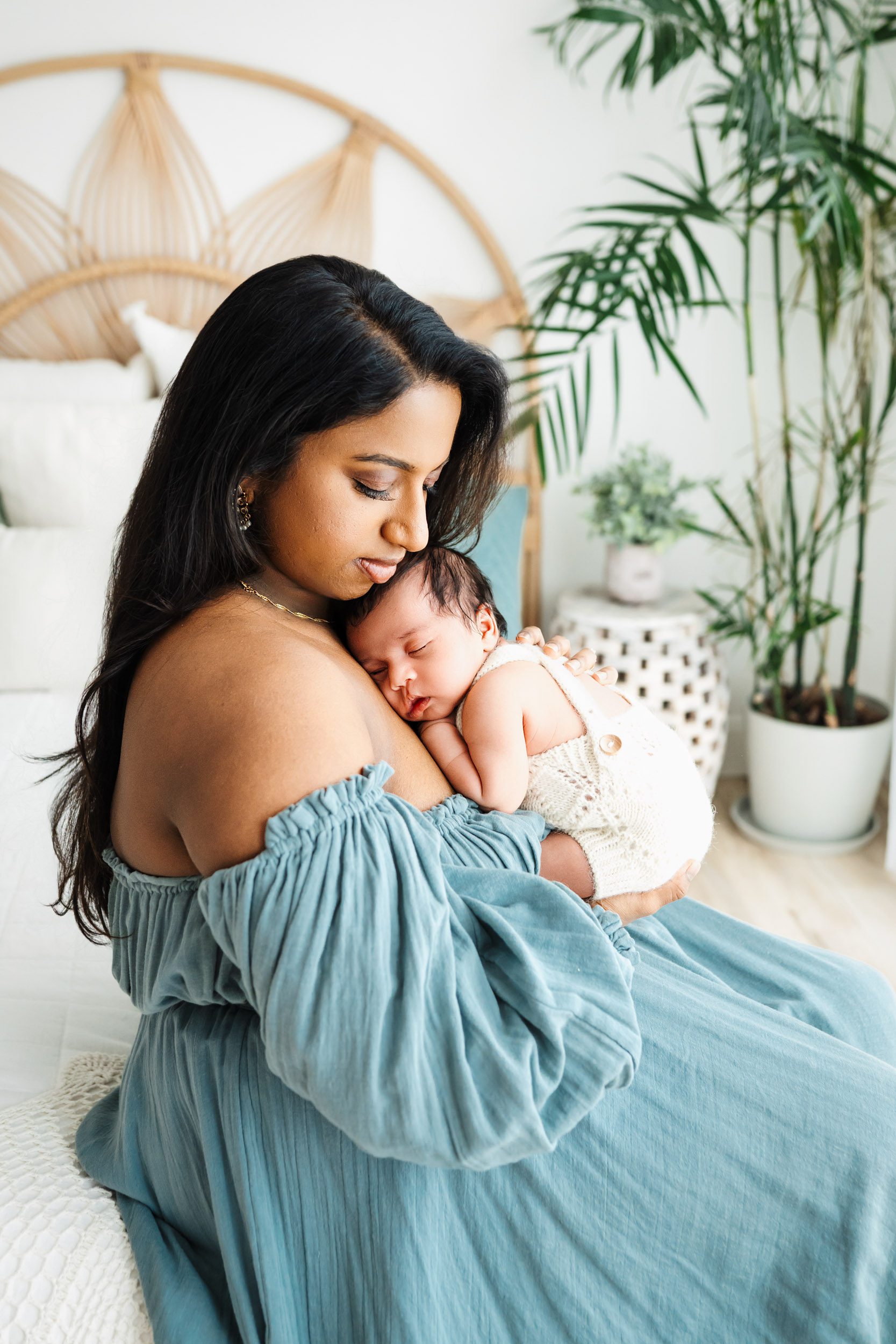 a new mom wearing a blue off the shoulder dress sitting on a bed with her baby boy snuggled up against her chest during a newborn photoshoot with mom