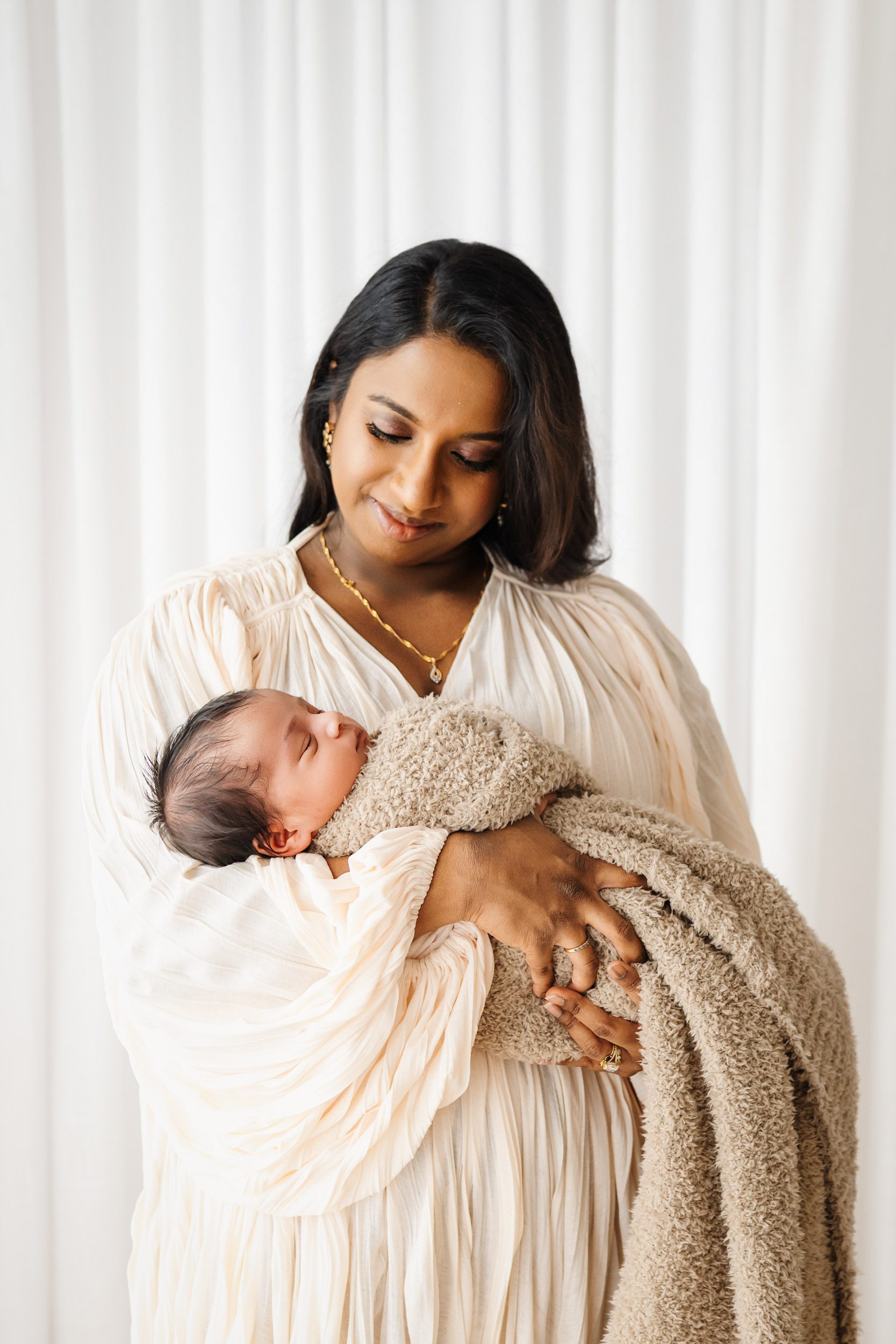 a new mom standing in front of a wall of white curtains cradling her baby boy in her arms as she smiles down at him during a newborn photoshoot with mom