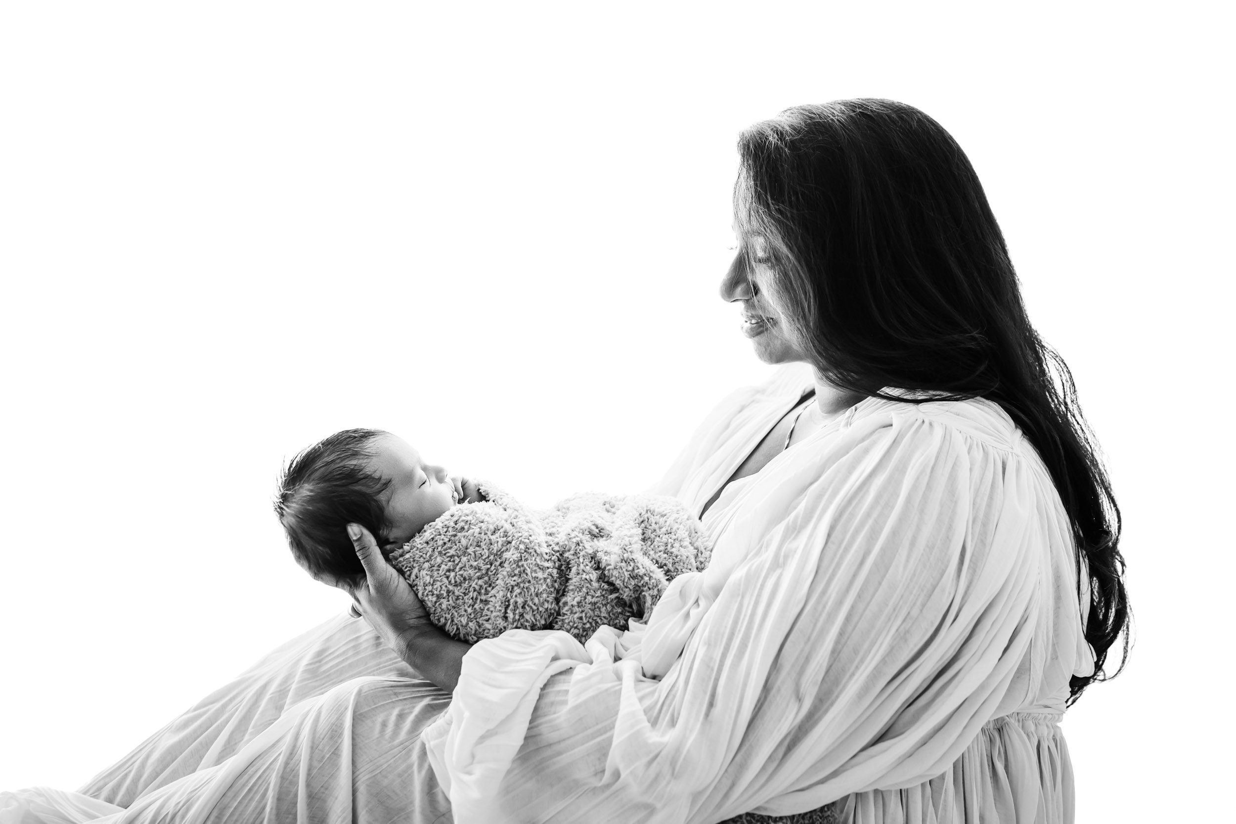 a black and white backlit photo of a new mom sitting and holding her baby boy as she smiles down at him a new mom wearing a blue off the shoulder dress sitting on a bed with her baby boy snuggled up against her chest during a newborn photoshoot with mom