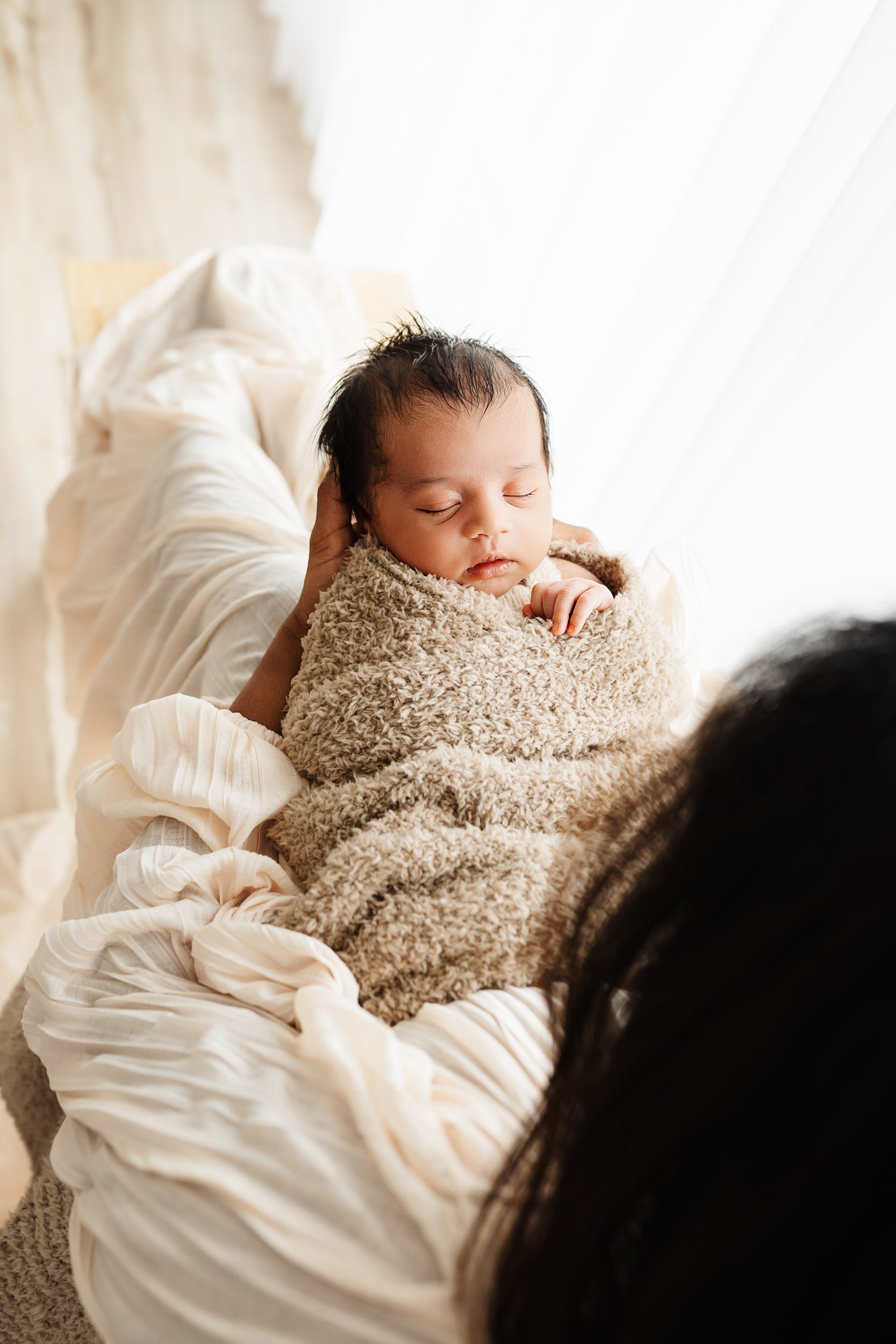 a photo taken from above of a baby boy wrapped in a taupe fuzzy blanket sleeping peacefully on his mom's legs as she looks down at him during a newborn photoshoot with mom