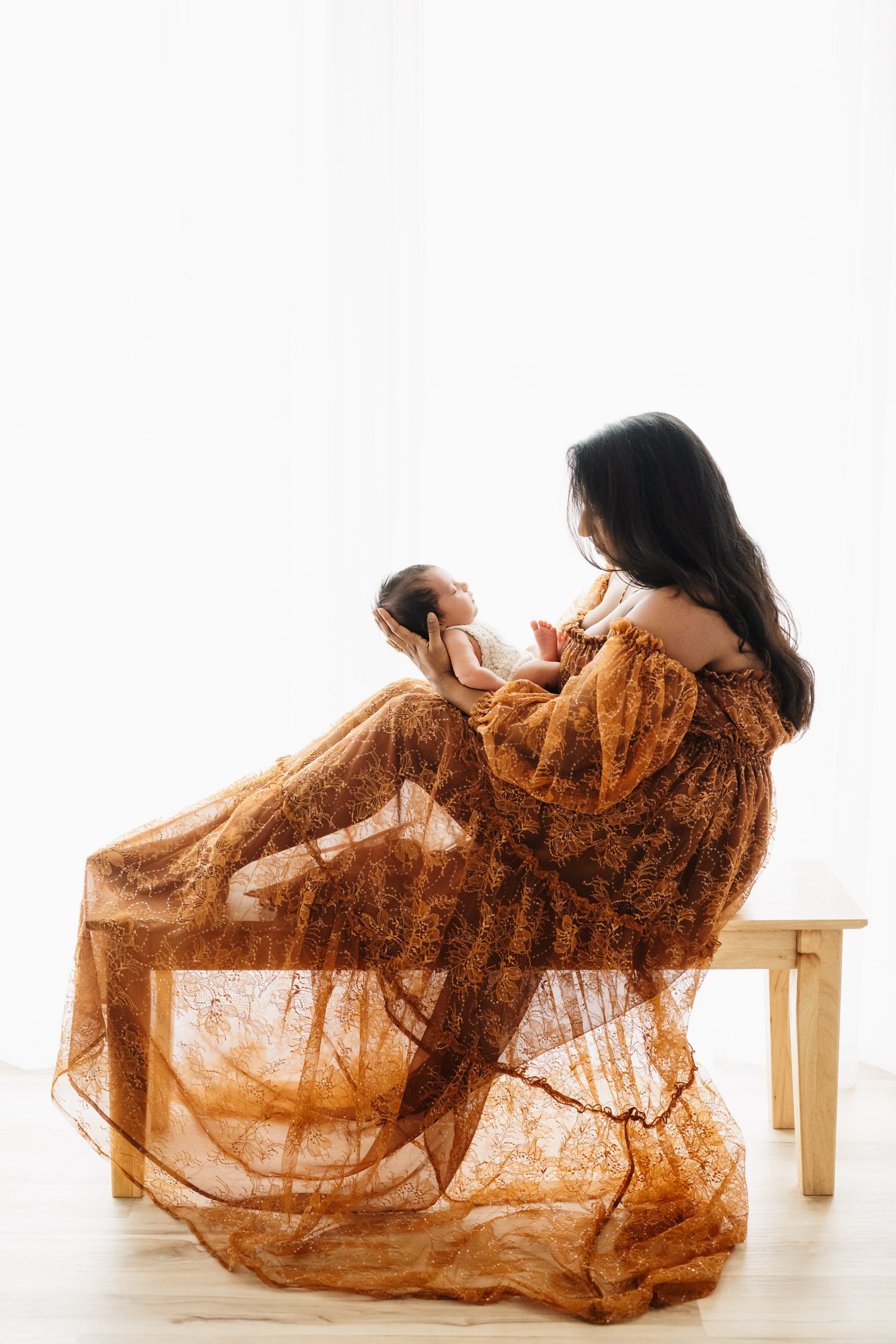 a backlit photo of a new mom wearing a rust colored lace dress sitting on a bench and holding her baby boy as she smiles down at him during a newborn photoshoot with mom