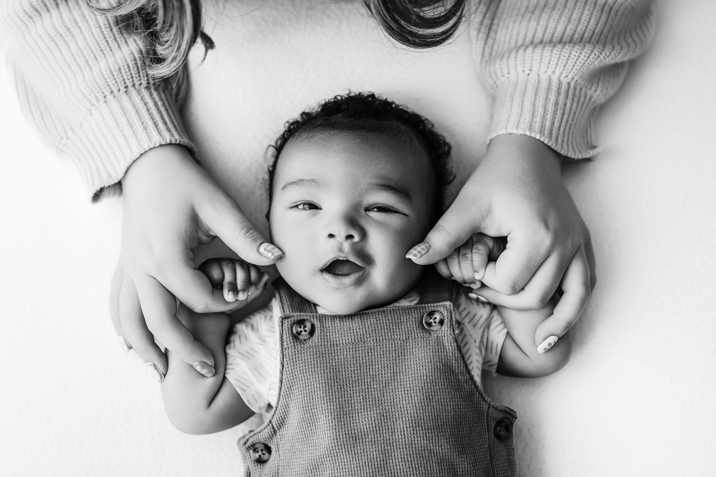a black and white picture of a baby boy holding onto his mom's fingers with his little hands and smiling up at the camera as she touches his chubby cheeks with her thumbs during a King of Prussia newborn photoshoot