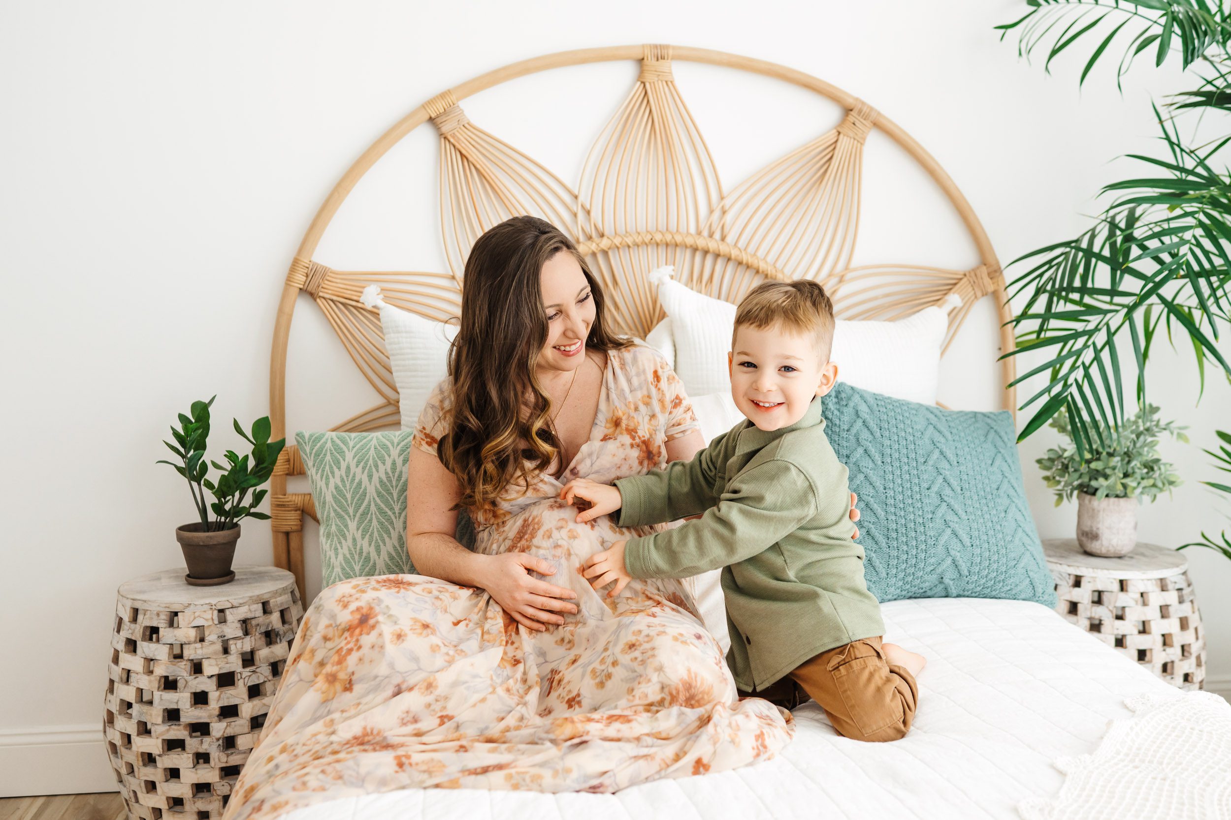 an expecting mom wearing a floral dress sitting on a bed and smiling at her young toddler as he tickles her belly and smiles at the camera during a maternity photoshoot