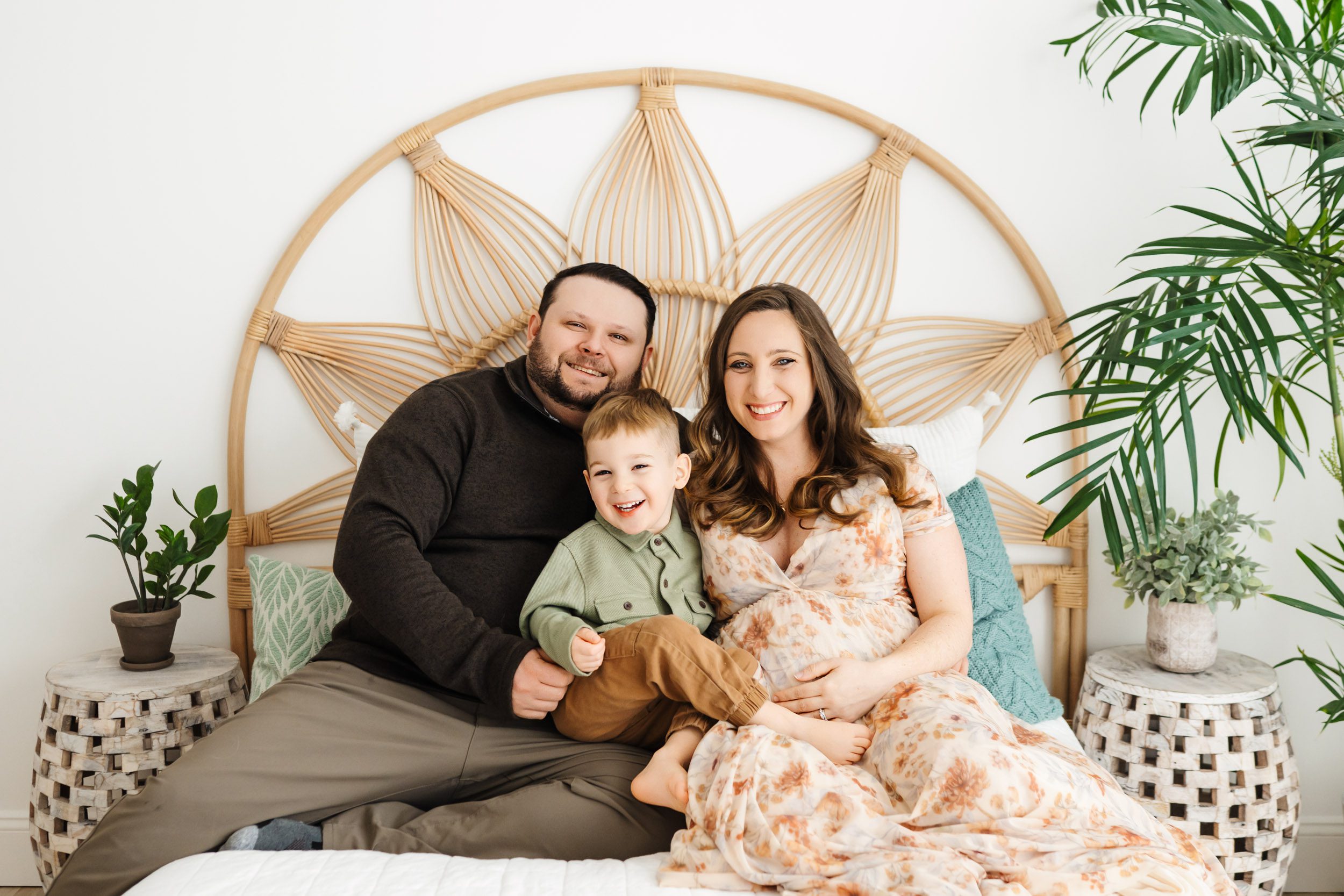 expecting parents sitting on a bed with their toddler on their lap as they all smile at the camera during a maternity photoshoot