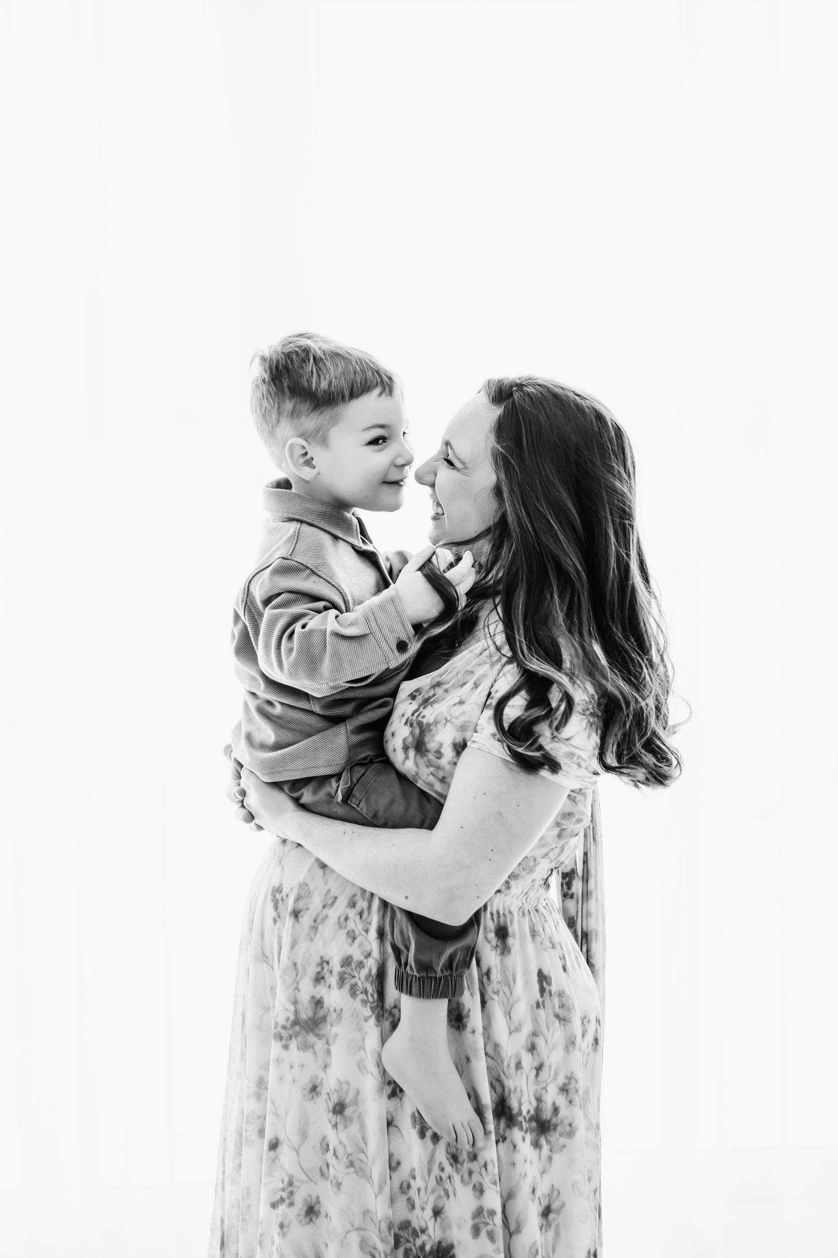 a black and white backlit photo of an expecting mom holding her toddler above her belly as they gently touch noses during a maternity photoshoot