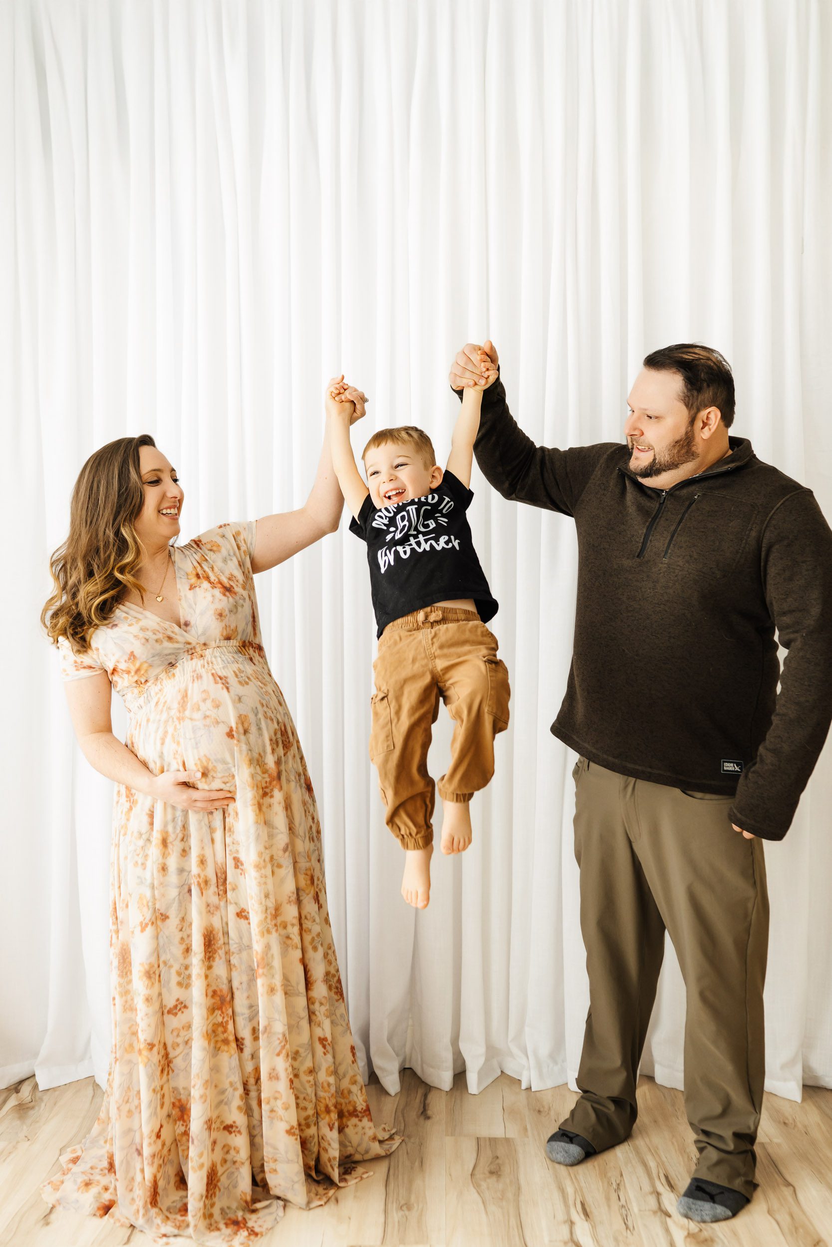 expecting parents holding their toddler's hands and lifting him up in the air as they all laugh during a maternity photoshoot