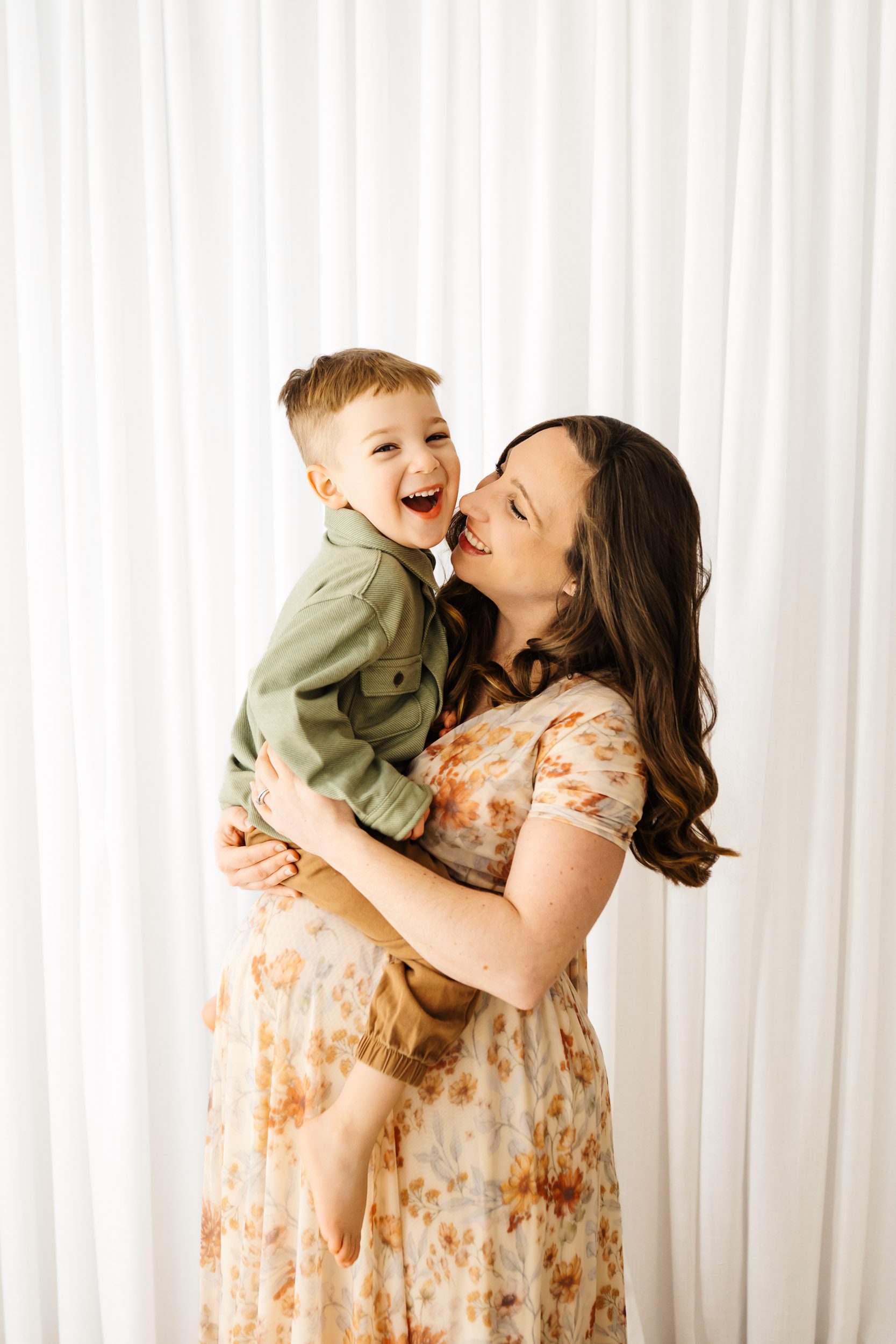 a pregnant mom wearing a floral dress holding her toddler above her belly and smiling at him as he looks at the camera and laughs during a maternity photoshoot