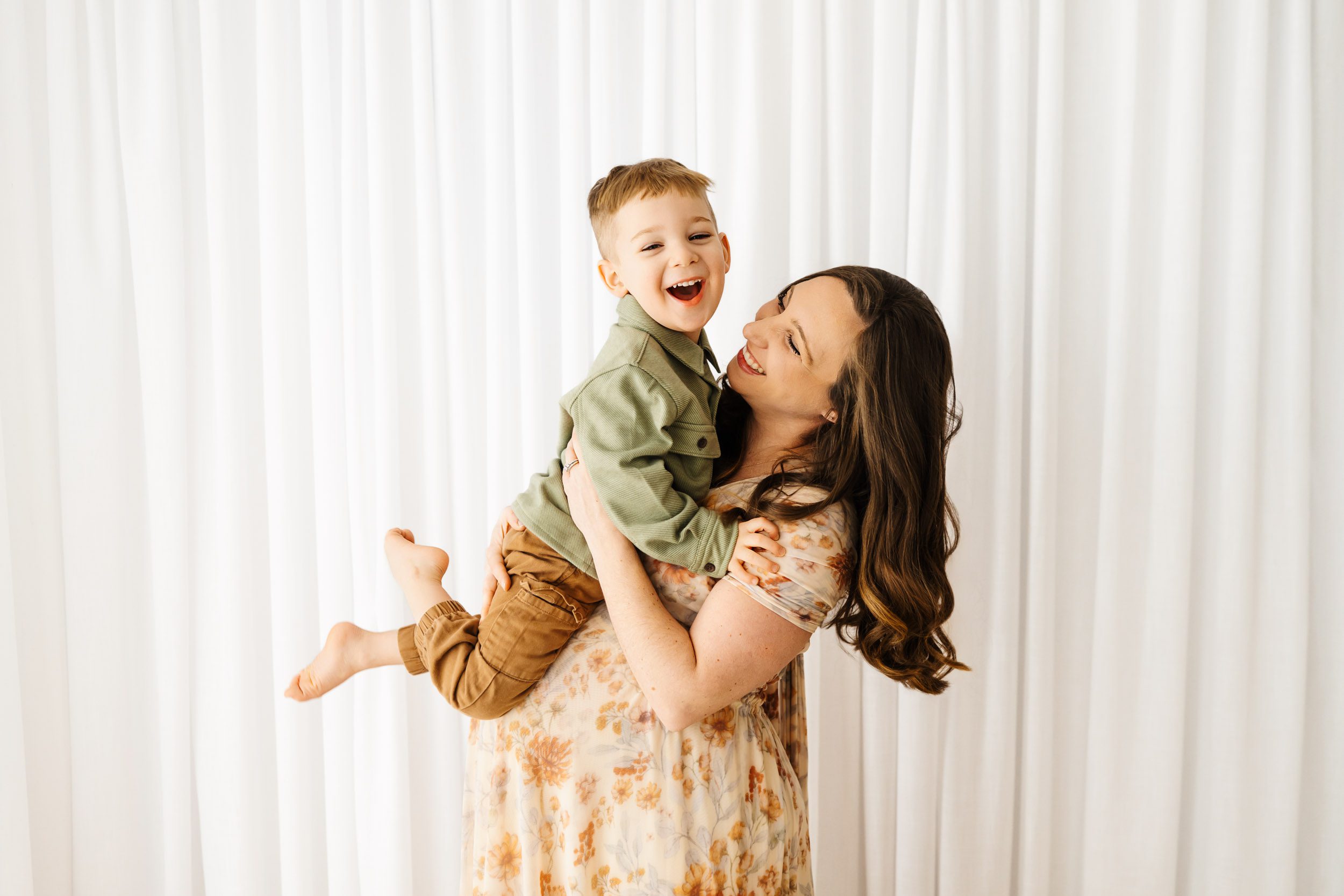a pregnant mom holding her toddler and lifting him up in the air as he smiles at the camera during a maternity photoshoot