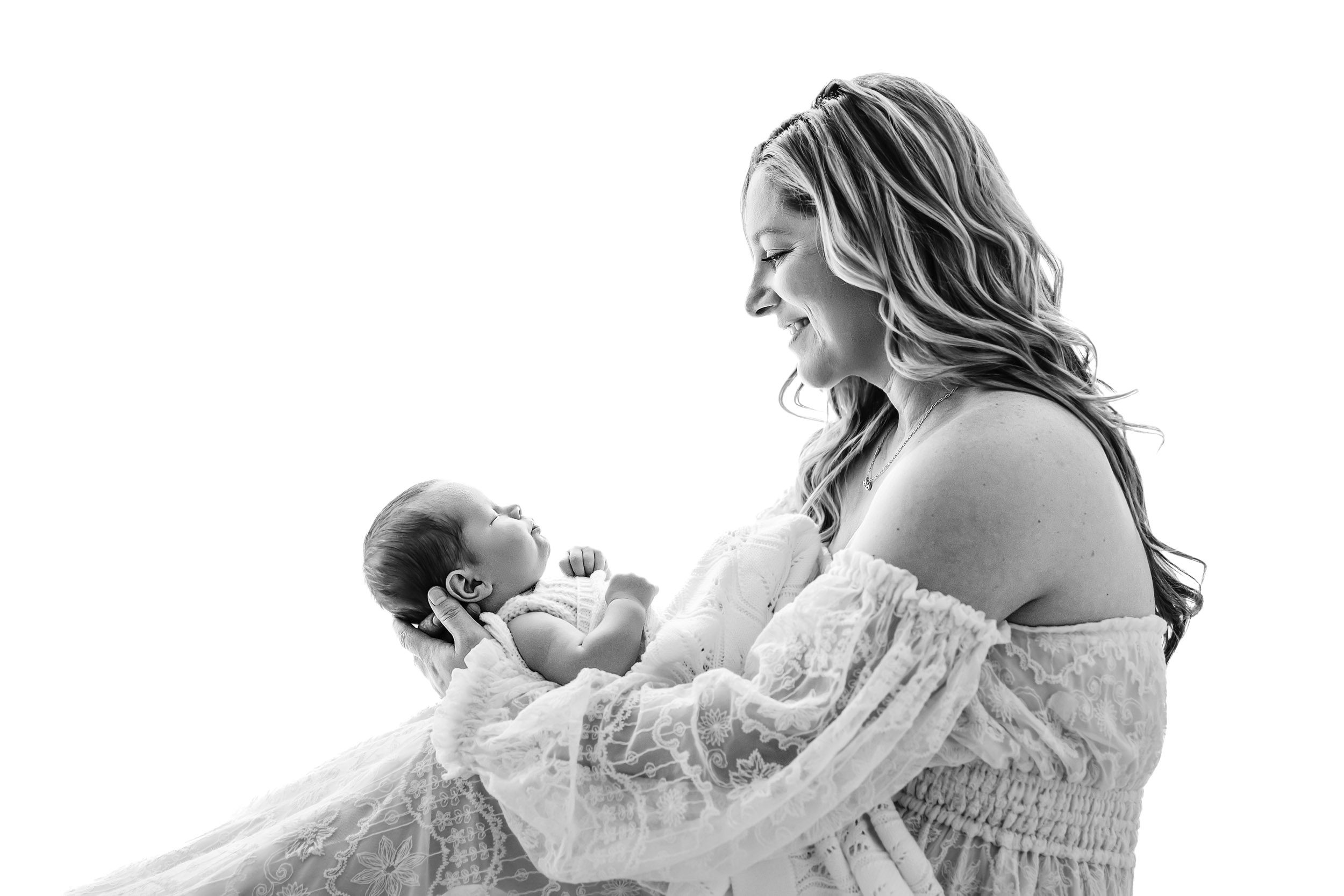 a black and white backlit photo of a new mother sitting on a bench holding her baby boy in her arms and smiling down at him during a family centered newborn photoshoot