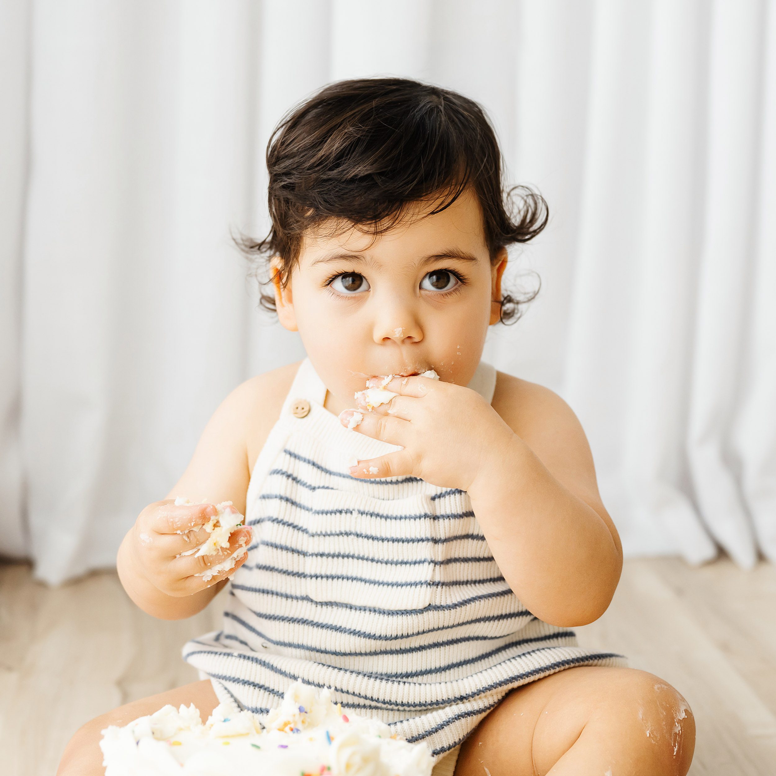 a close up picture of a little boy sitting on the floor in front of his birthday cake and gazing up toward the camera as he puts a piece of cake in his mouth