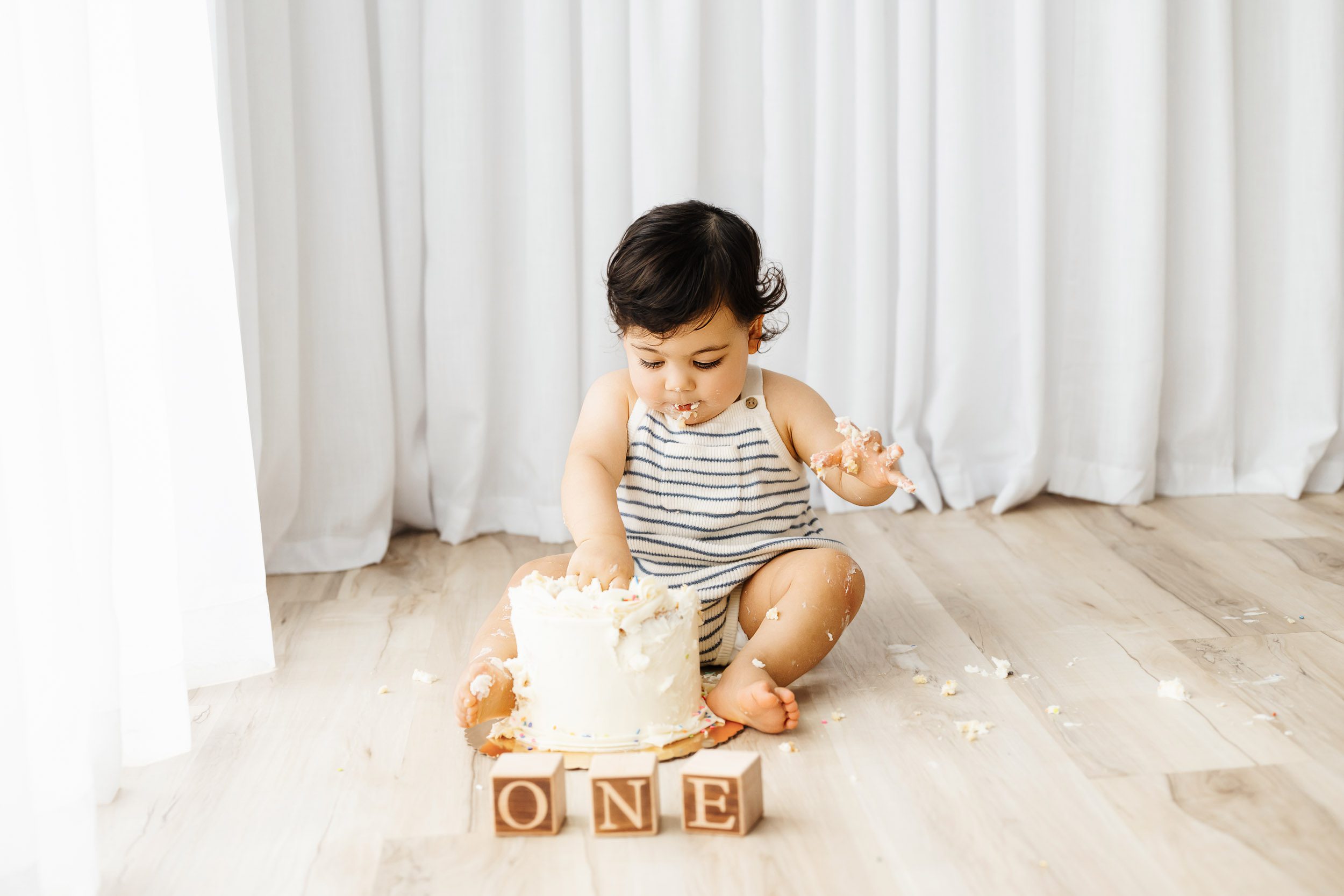 a baby boy covered in icing sitting on the floor squishing his hands into his birthday cake during a cake smash photoshoot