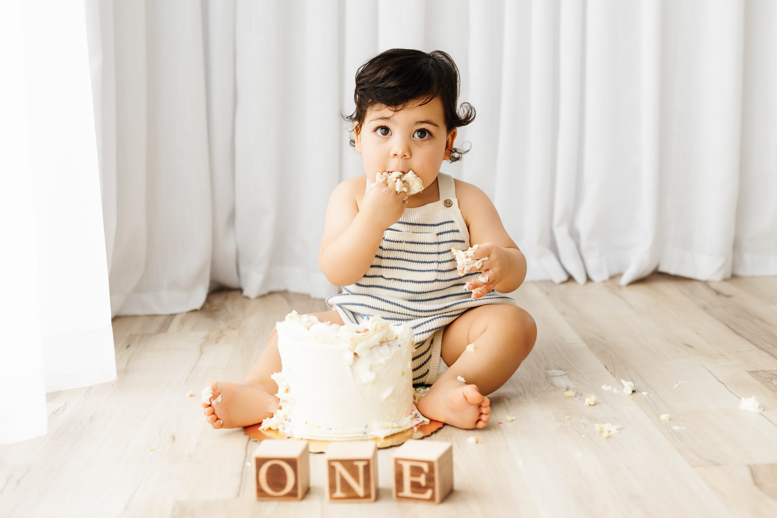 a little boy sitting on the floor in front of a wall of white curtains gazing at the camera as he puts a fist full of cake and icing in his mouth during a cake smash photoshoot